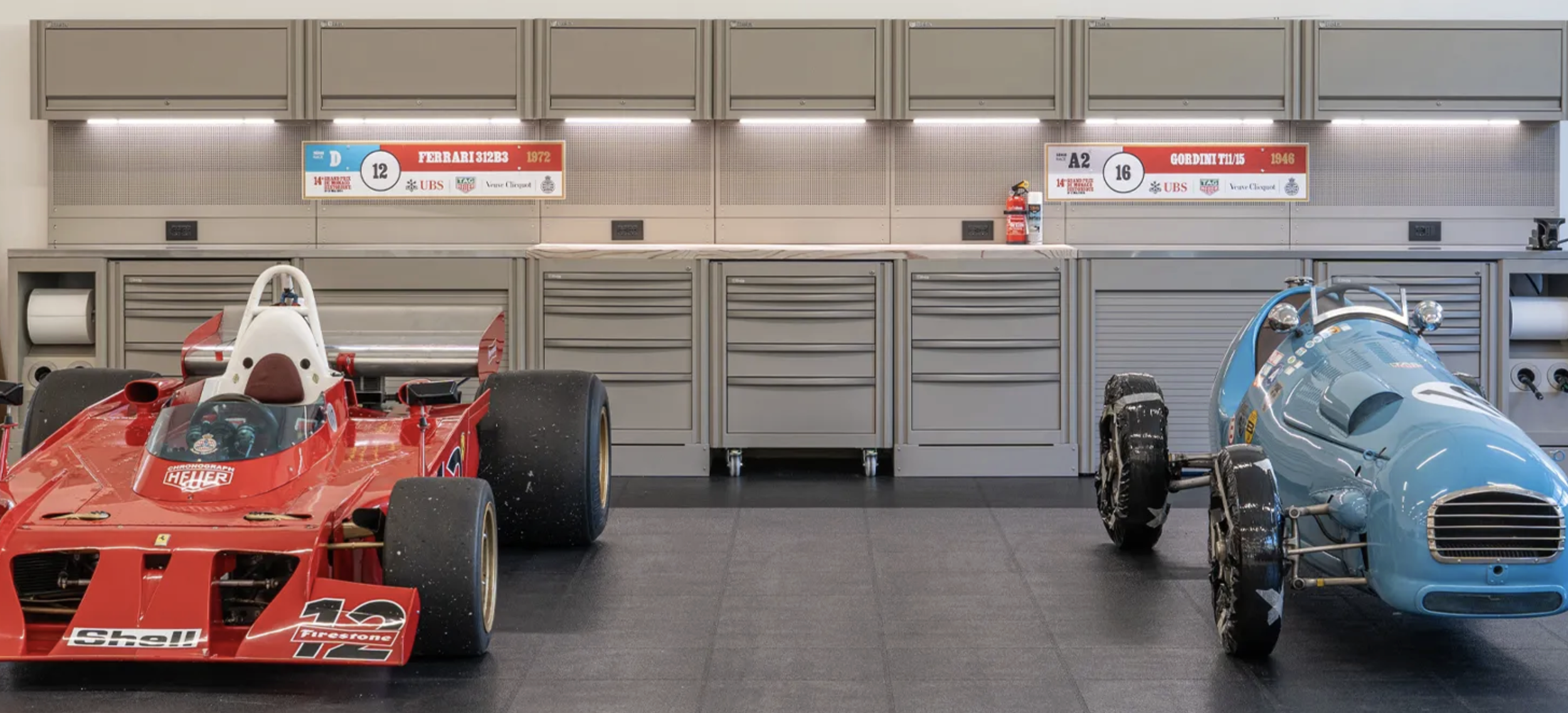 To Formel 1-biler i et garasjemiljø, en rød Ferrari og en blå Williams, med verktøy og utstyr på bakveggen.