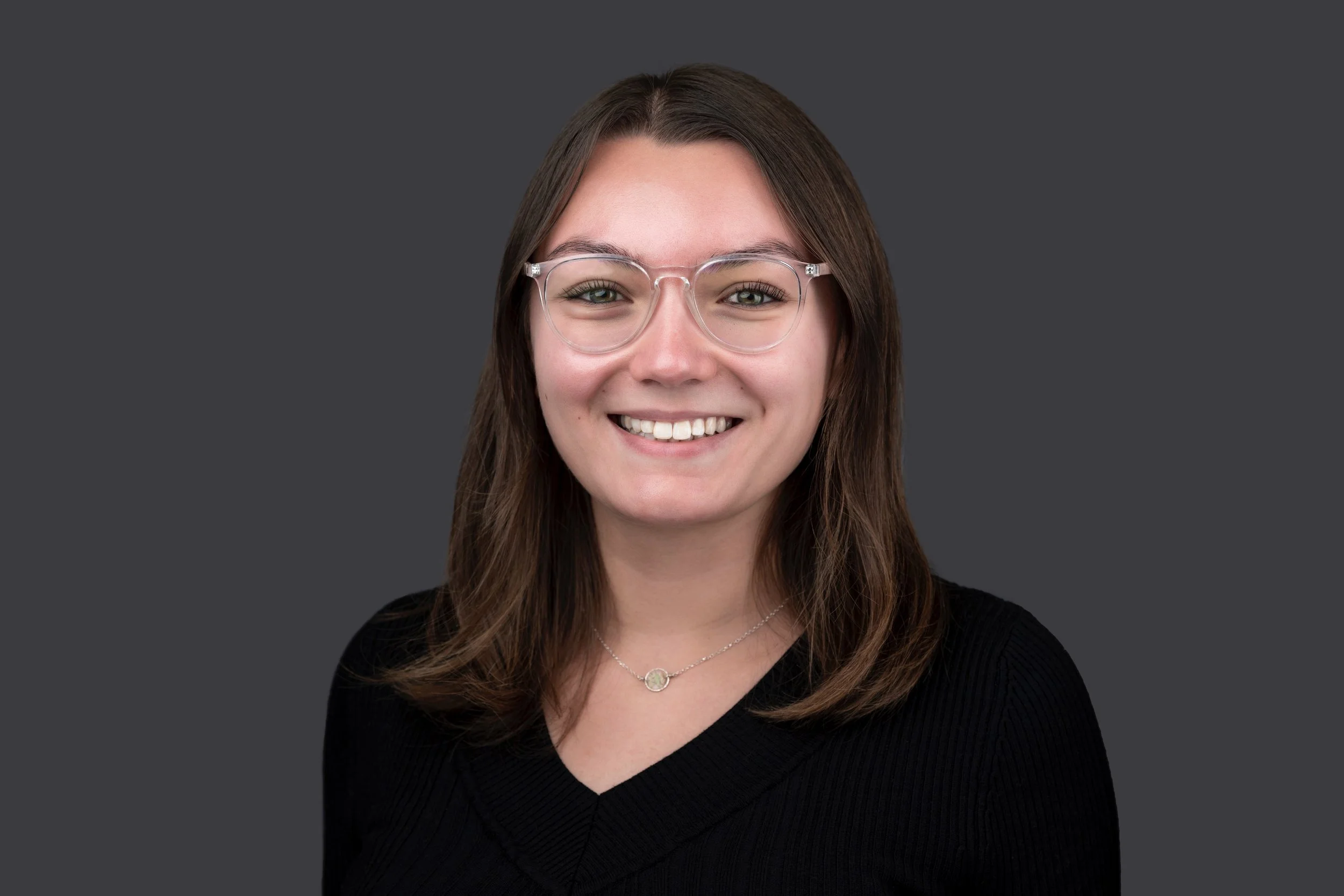 A young woman with shoulder-length brown hair, wearing clear glasses and a black top, smiling against a dark gray background.