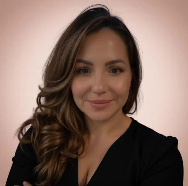 Close-up portrait of a woman with long wavy brown hair, smiling, wearing a black top, against a plain light pink background.