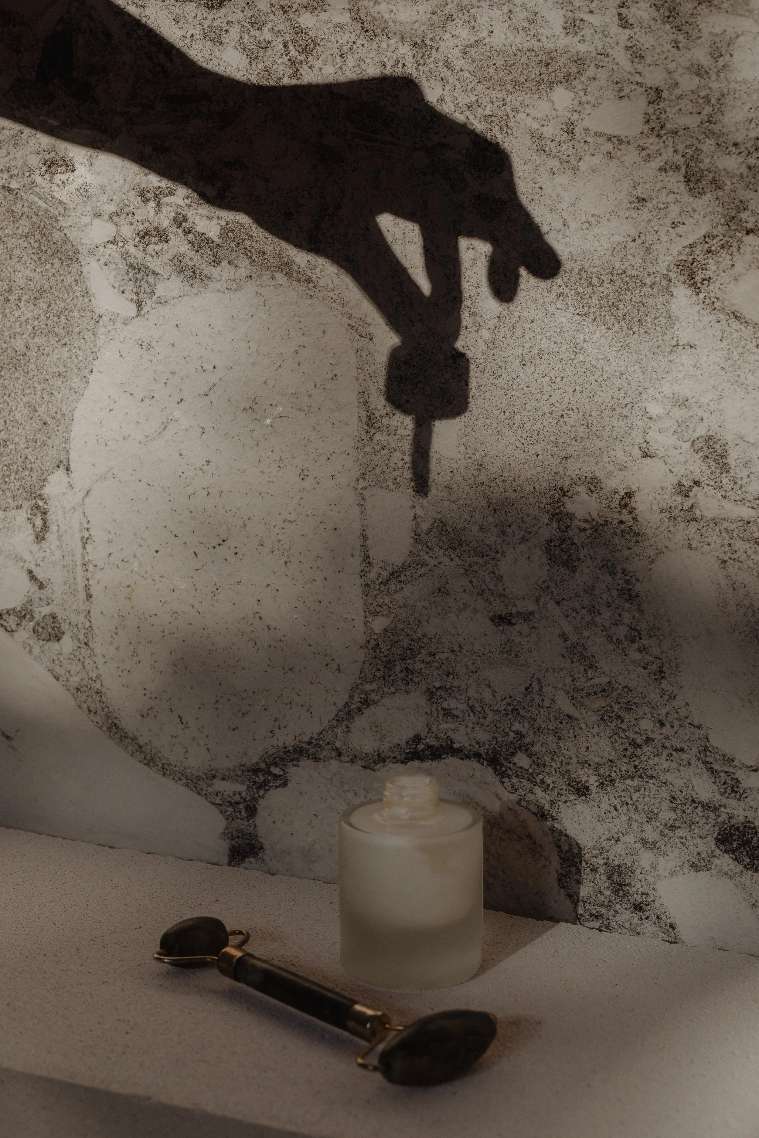 A shadow of a hand holding a small object casts on a textured surface with a white jar, a pair of dark glasses, and a black tool in the foreground.