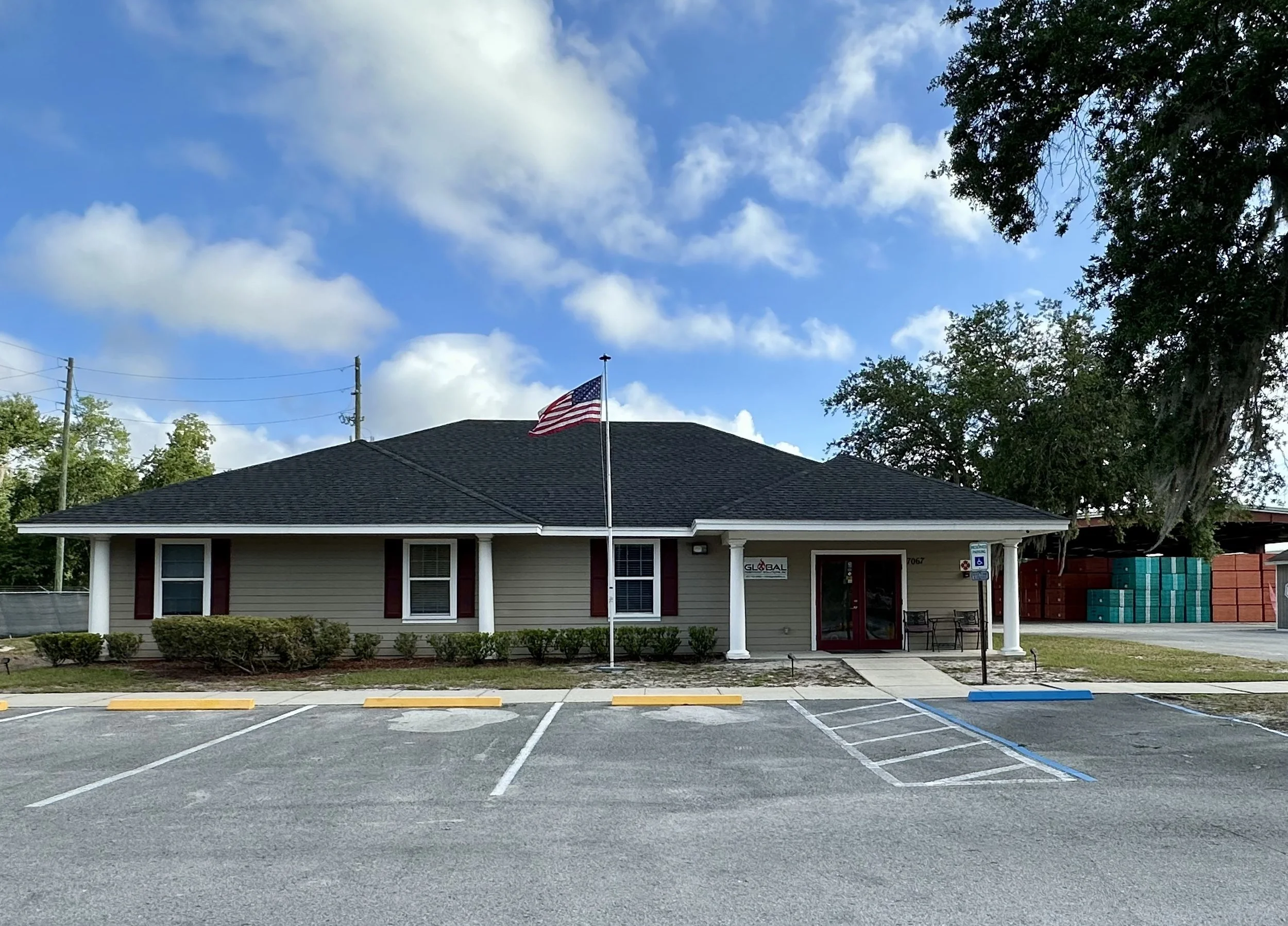 A building with a dark gray roof, white columns, and beige siding, with an American flag flying on a flagpole in front. There are shrubs and a parking lot with yellow and blue marked spaces, and trees with green leaves. The sky is blue with scattered white clouds.