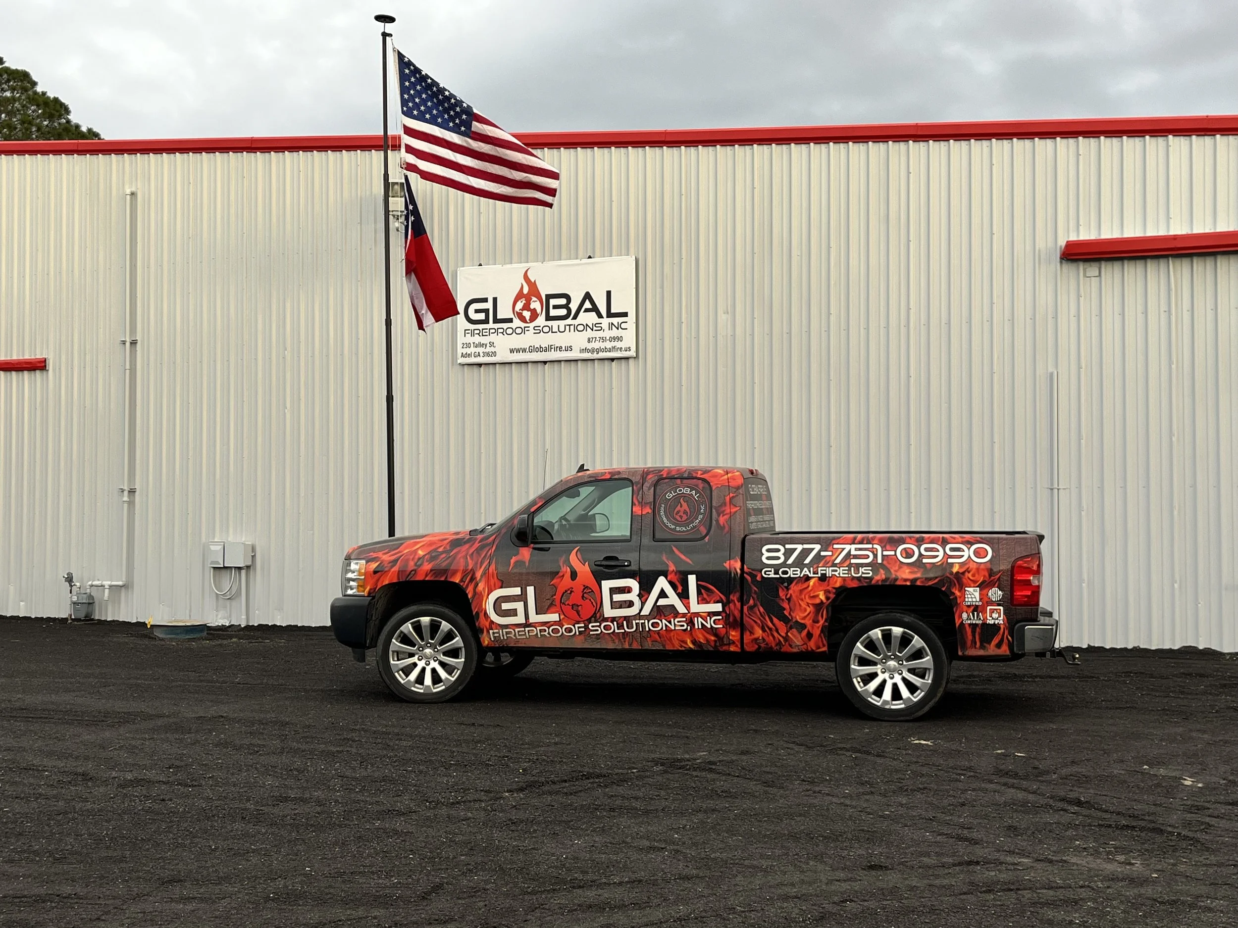 A red pickup truck with flame graphics and the company logo parked in front of a commercial building. The building has beige metal siding with red accents on the roof and edges. Two flags, the American flag and a red flag, are flying on poles in front of the building. There's a sign on the building that reads 'Global Fireproof Solutions, Inc.' along with website and contact information.