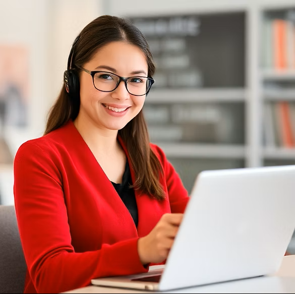 Woman wearing glasses and a red sweater, sitting at a desk with a laptop, smiling, in an office setting.