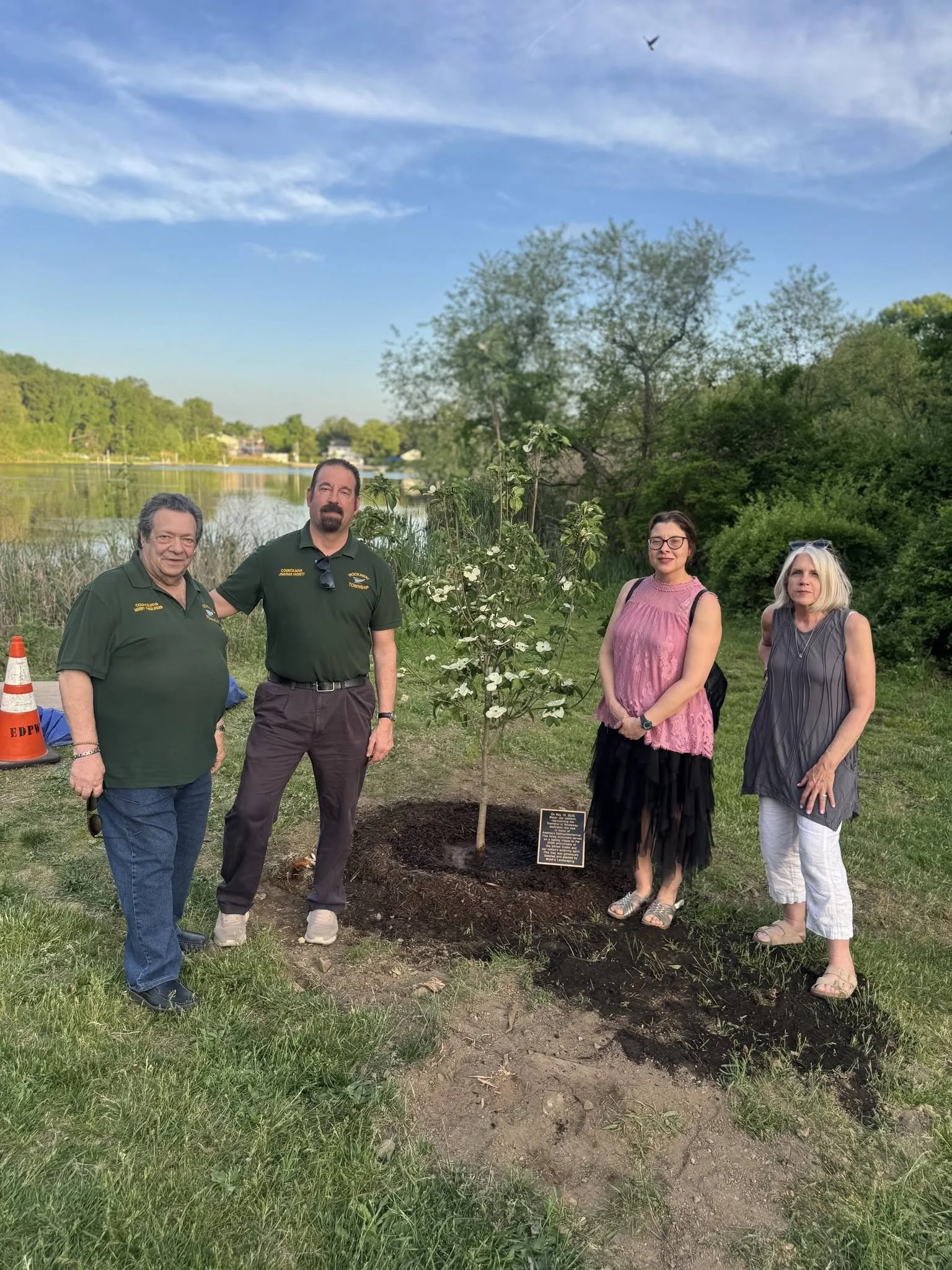Four people standing next to a newly planted tree near a river, with trees, water, and a blue sky in the background.