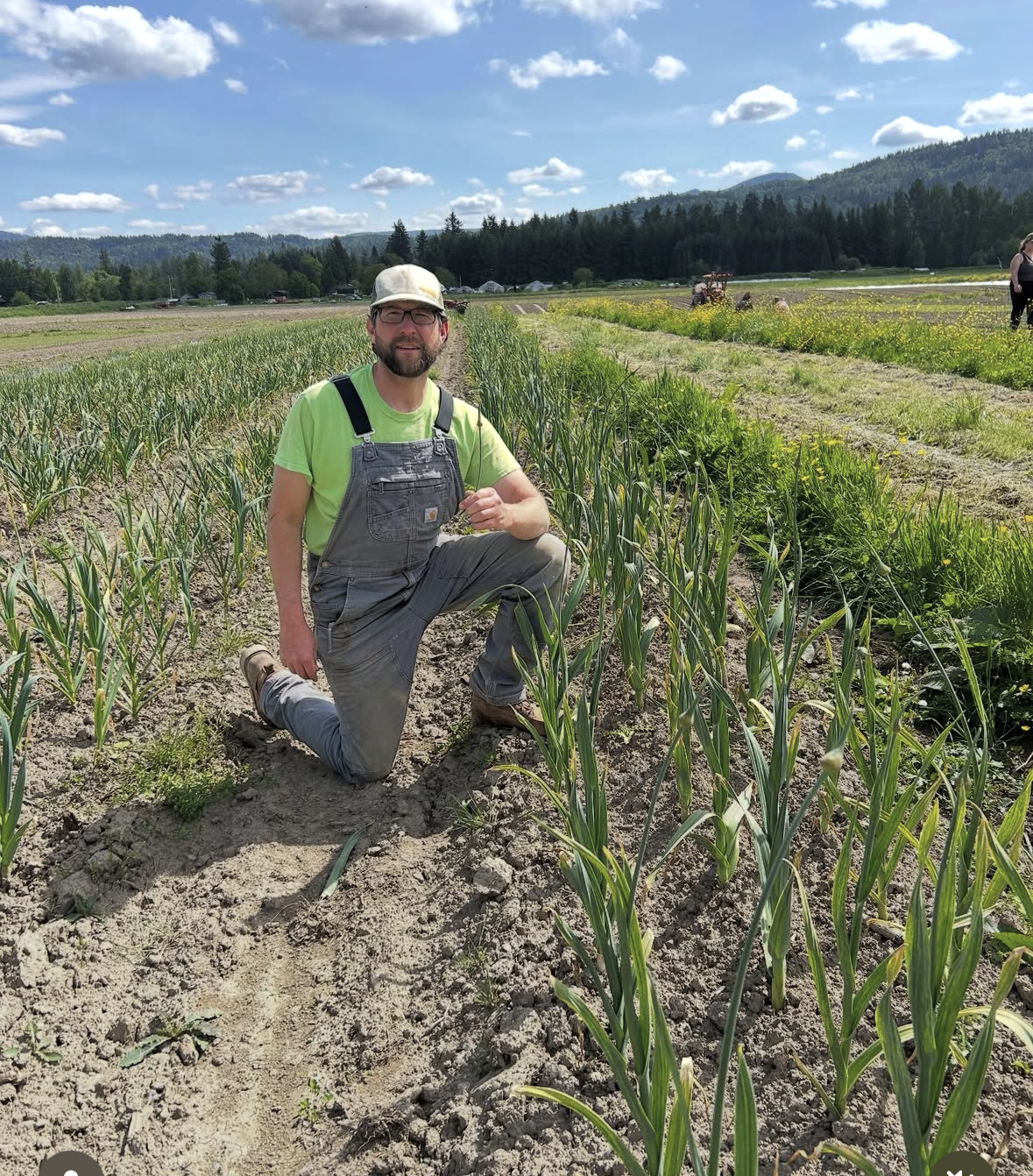 A man kneeling in a field of crops with rolling hills and a blue sky with scattered clouds in the background.