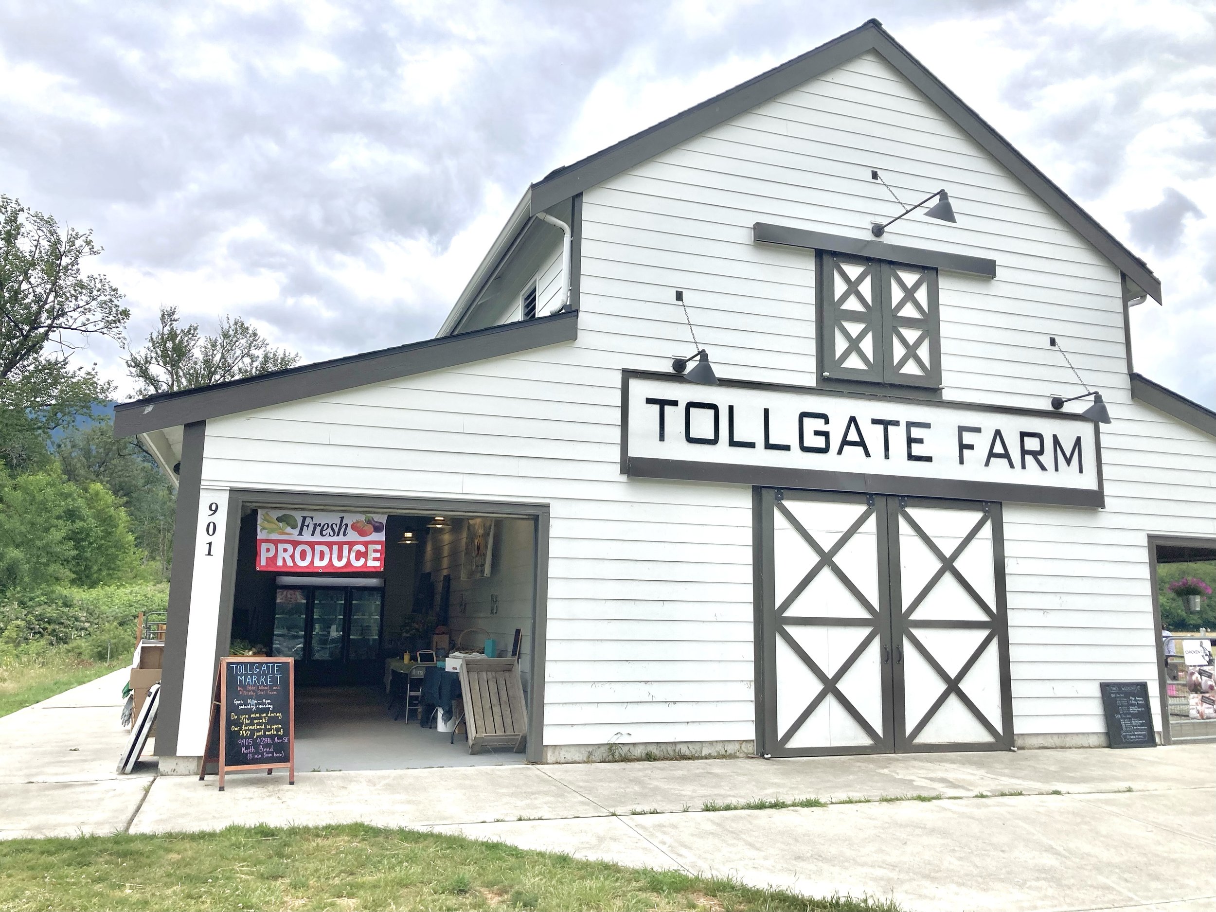 White farm building with large sign reading 'Tollgate Farm,' an open entrance with a sign advertising fresh produce, and black barn-style double doors.
