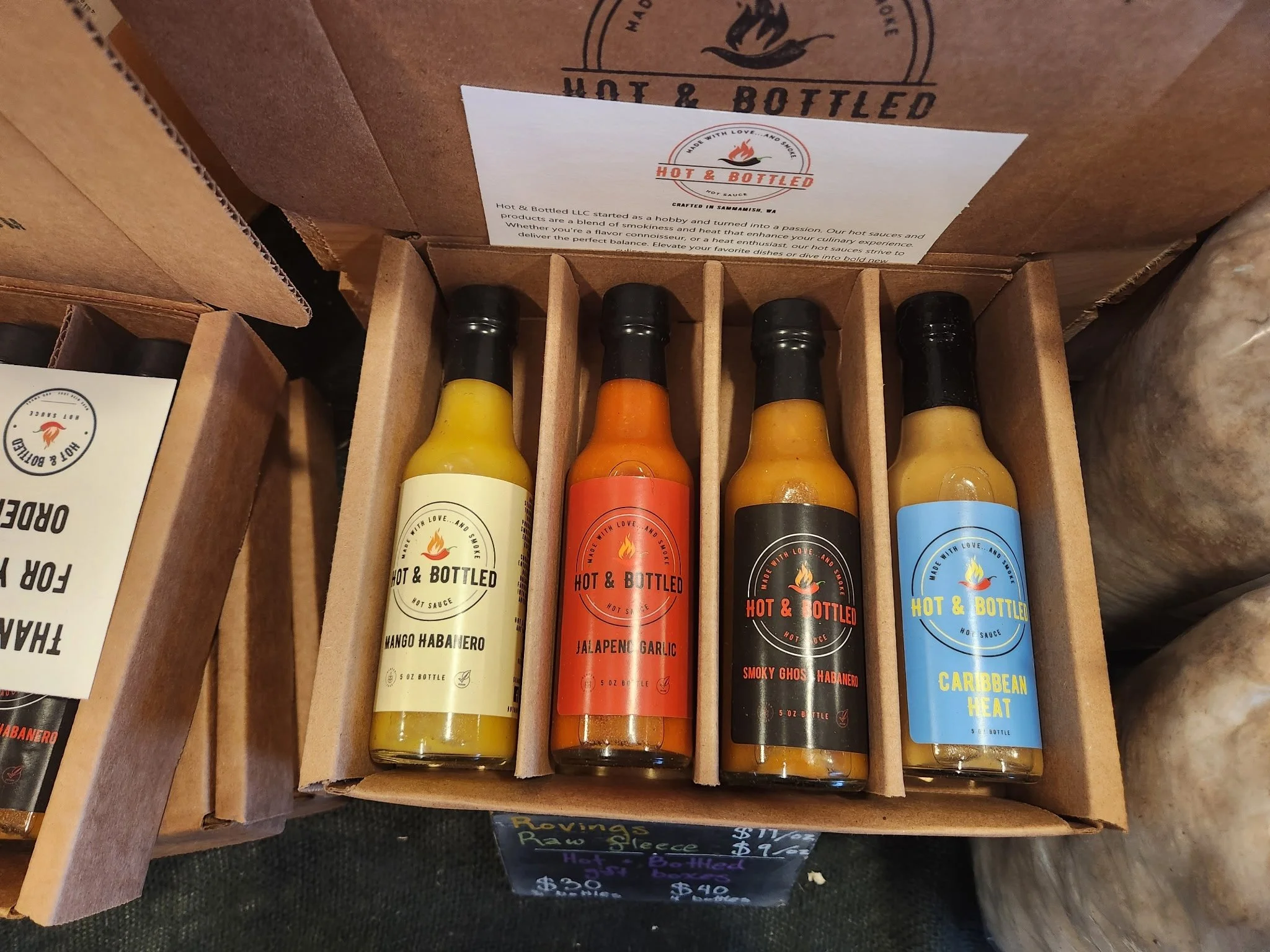 Four bottles of hot sauce from Hot & Bottled brand, arranged inside a cardboard box. The flavors from left to right are Mango Habanero, Jalapeno Garlic, Smoky Gho Habanero, and Caribbean Heat.