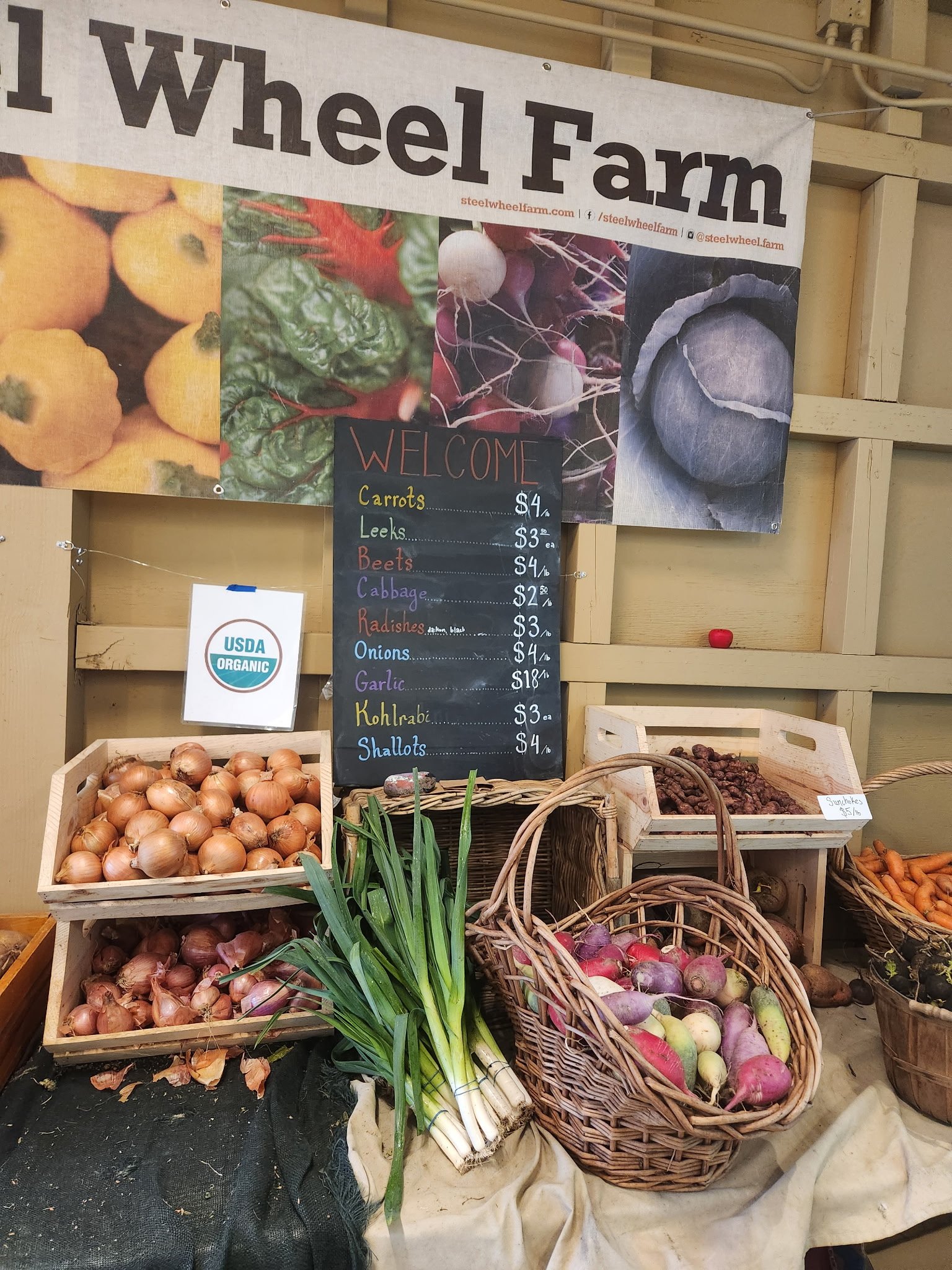 Display of fresh vegetables at a farm stand including onions, spring onions, beets, radishes, and turnips, with a blackboard showing prices and an 'USDA Organic' sign.