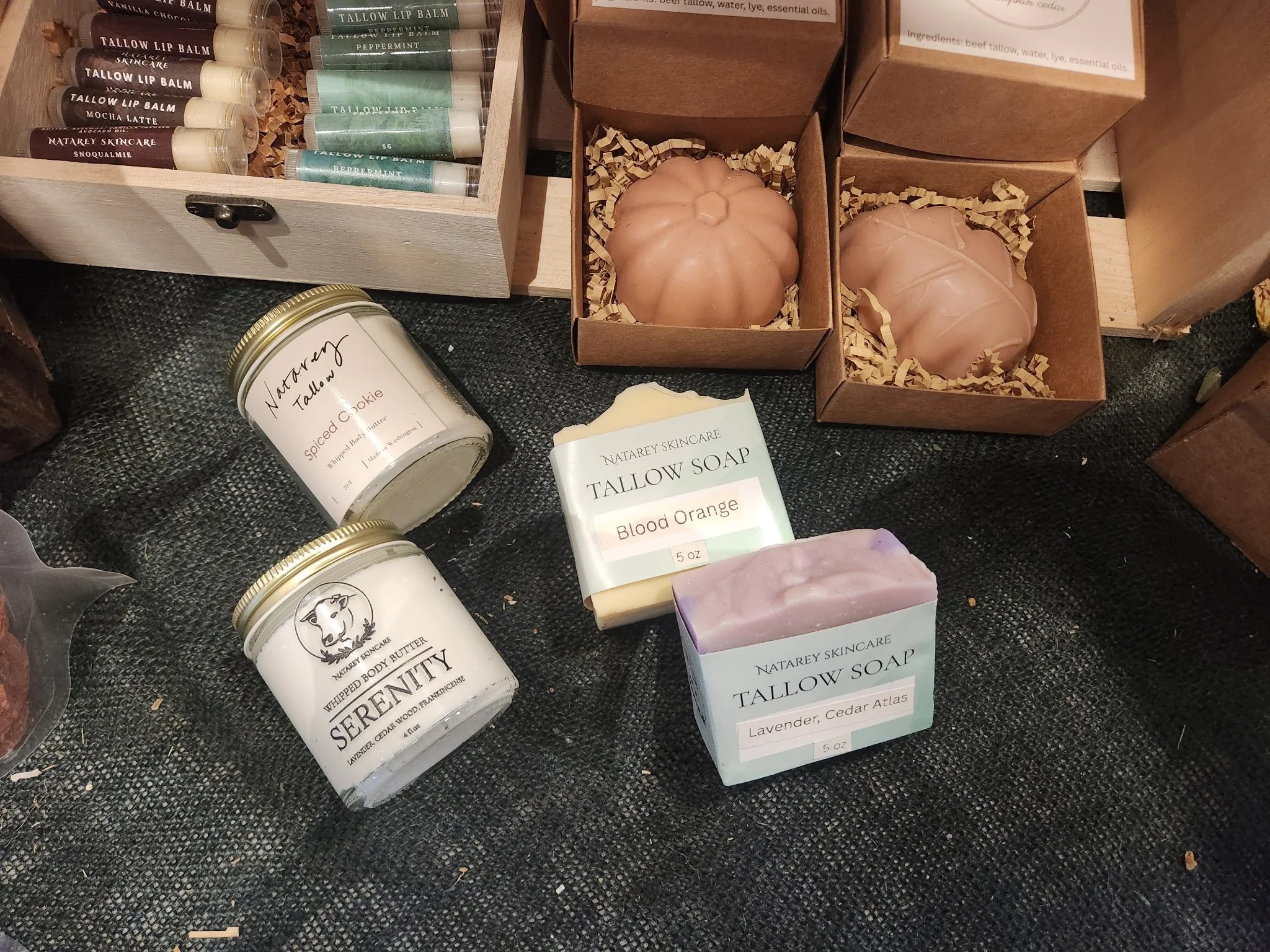 Several handmade soaps, including lavender and cedar atlas scent, and blood orange scent, displayed on a black fabric surface along with some pumpkin-shaped bath bombs and lip balms in containers.