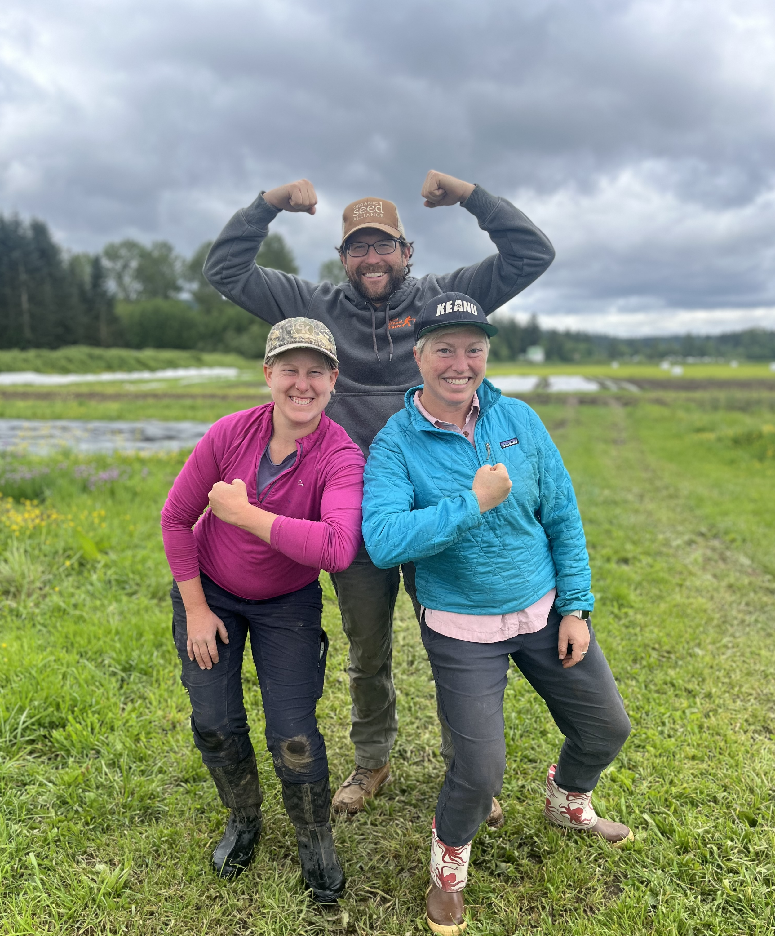 Three people outdoors on a farm, flexing and smiling, with fields and cloudy sky in the background.