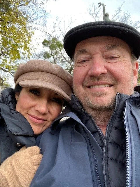 A man and woman smiling outdoors, wearing hats and jackets, with trees in the background.