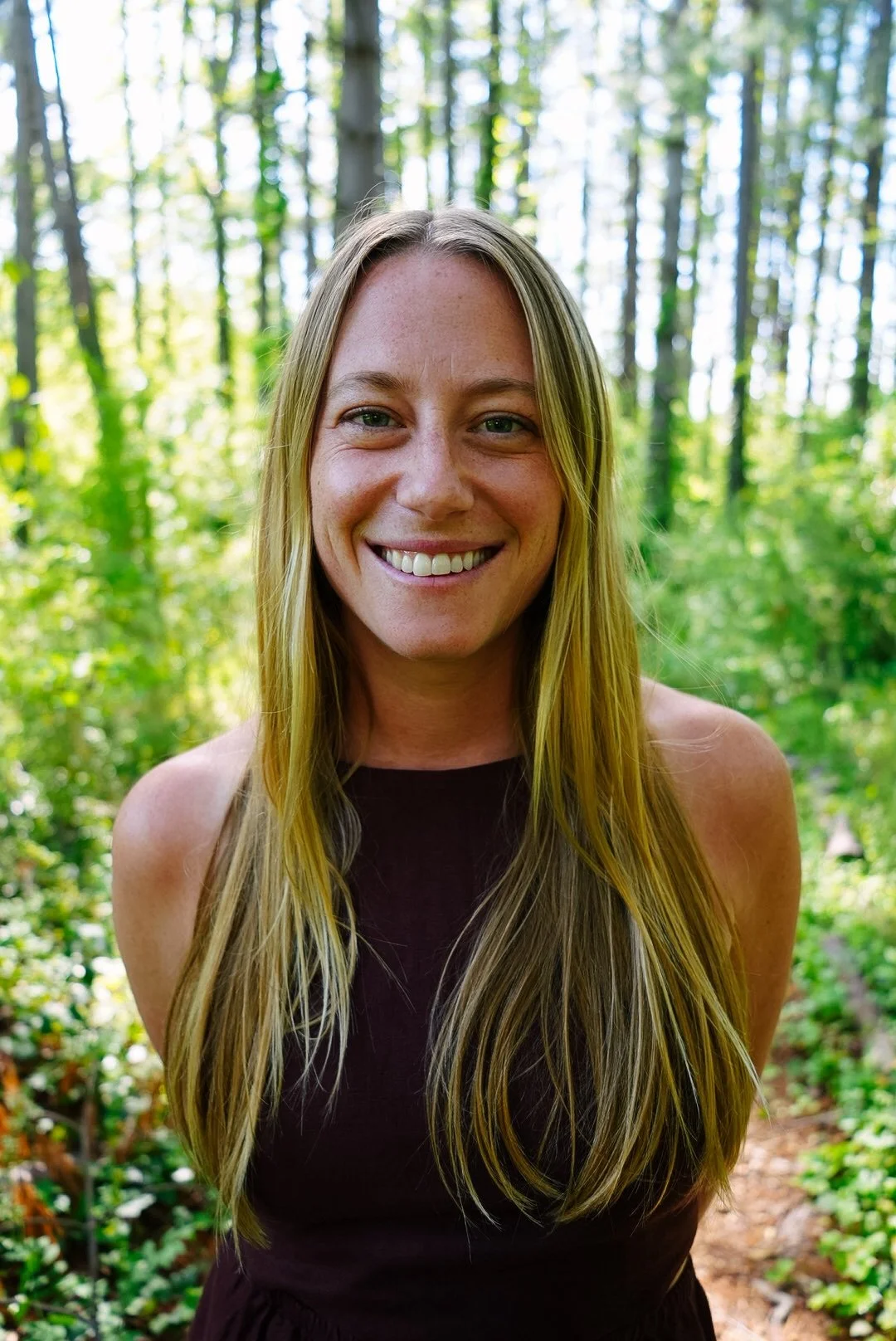A woman with long blonde hair smiling in a forest with green trees and foliage in the background.