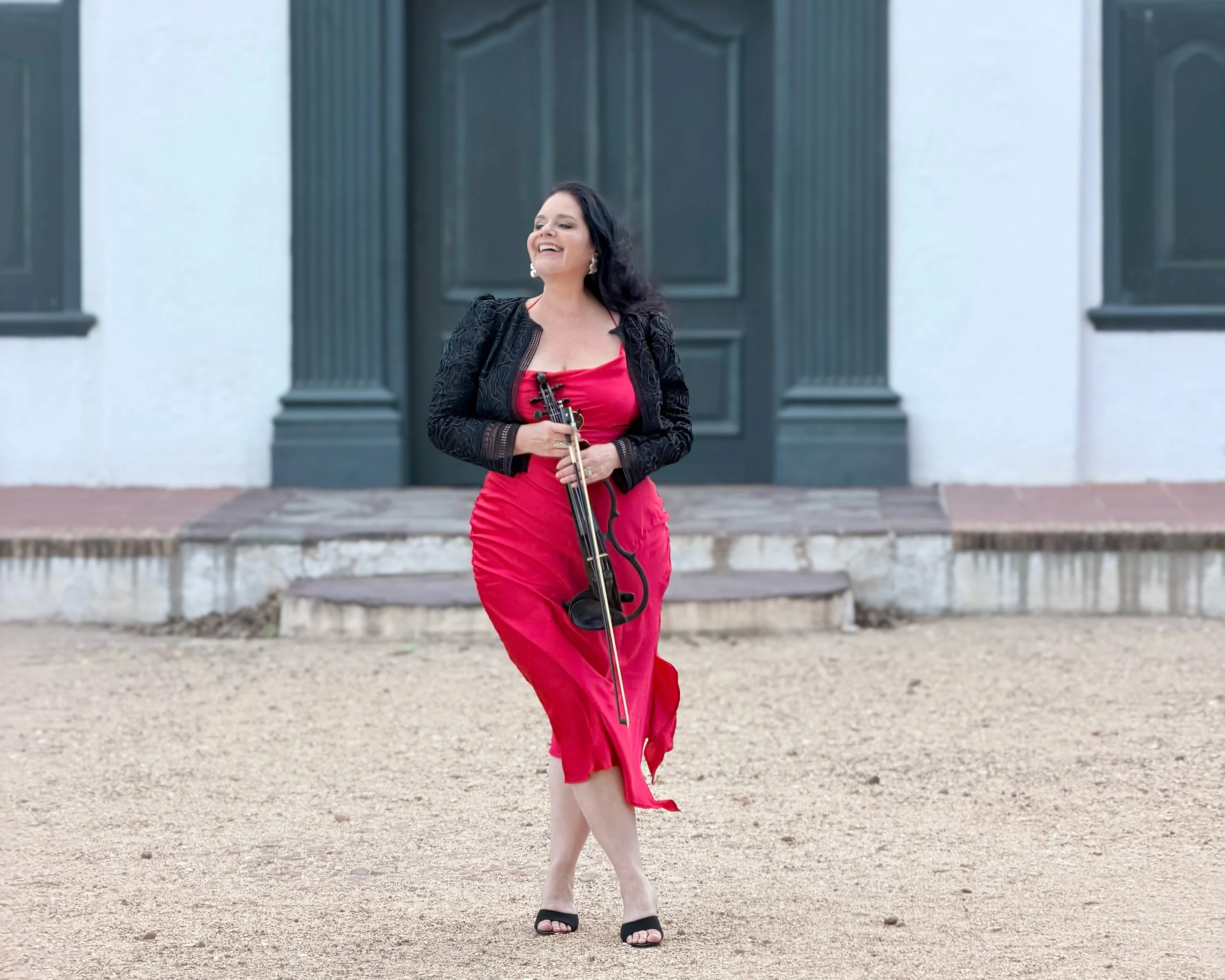 A woman in a red dress and black jacket holding a violin and bow, smiling and walking outside near a building with dark green doors.