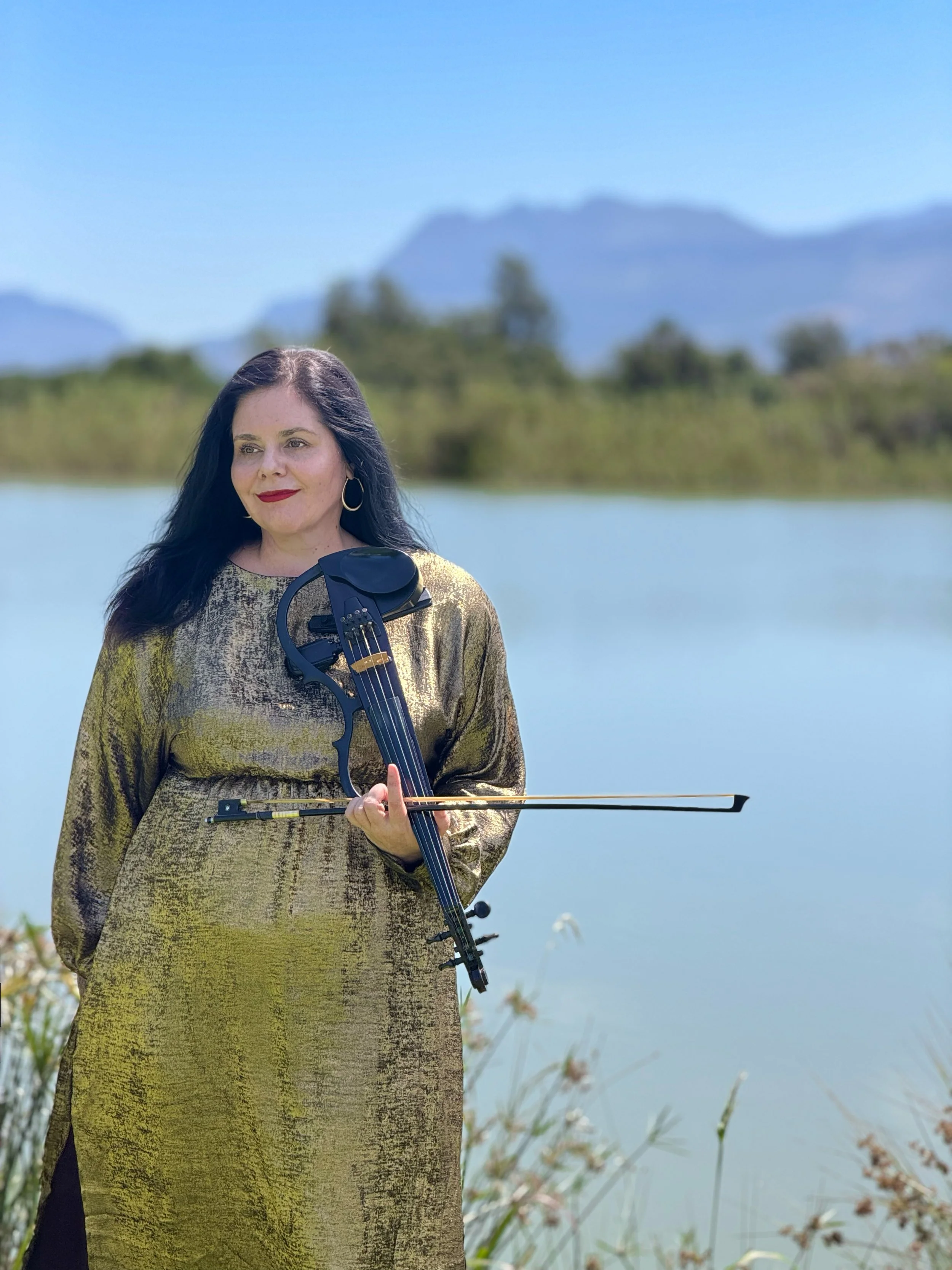A woman in a pink dress and orange jacket holding a violin and bow outdoors on a sunny day, with a large white umbrella behind her.
