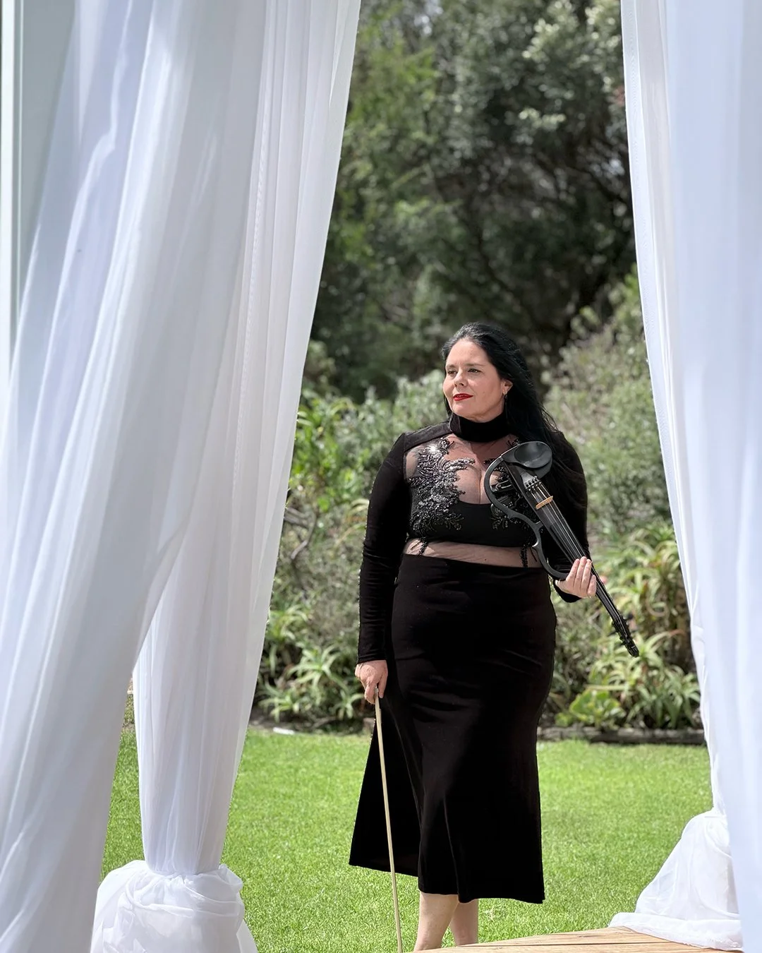 A woman with black hair, wearing a black dress with sheer and embroidered details, holding a violin in one hand and a bow in the other, walking outdoors through white curtains, with greenery in the background.