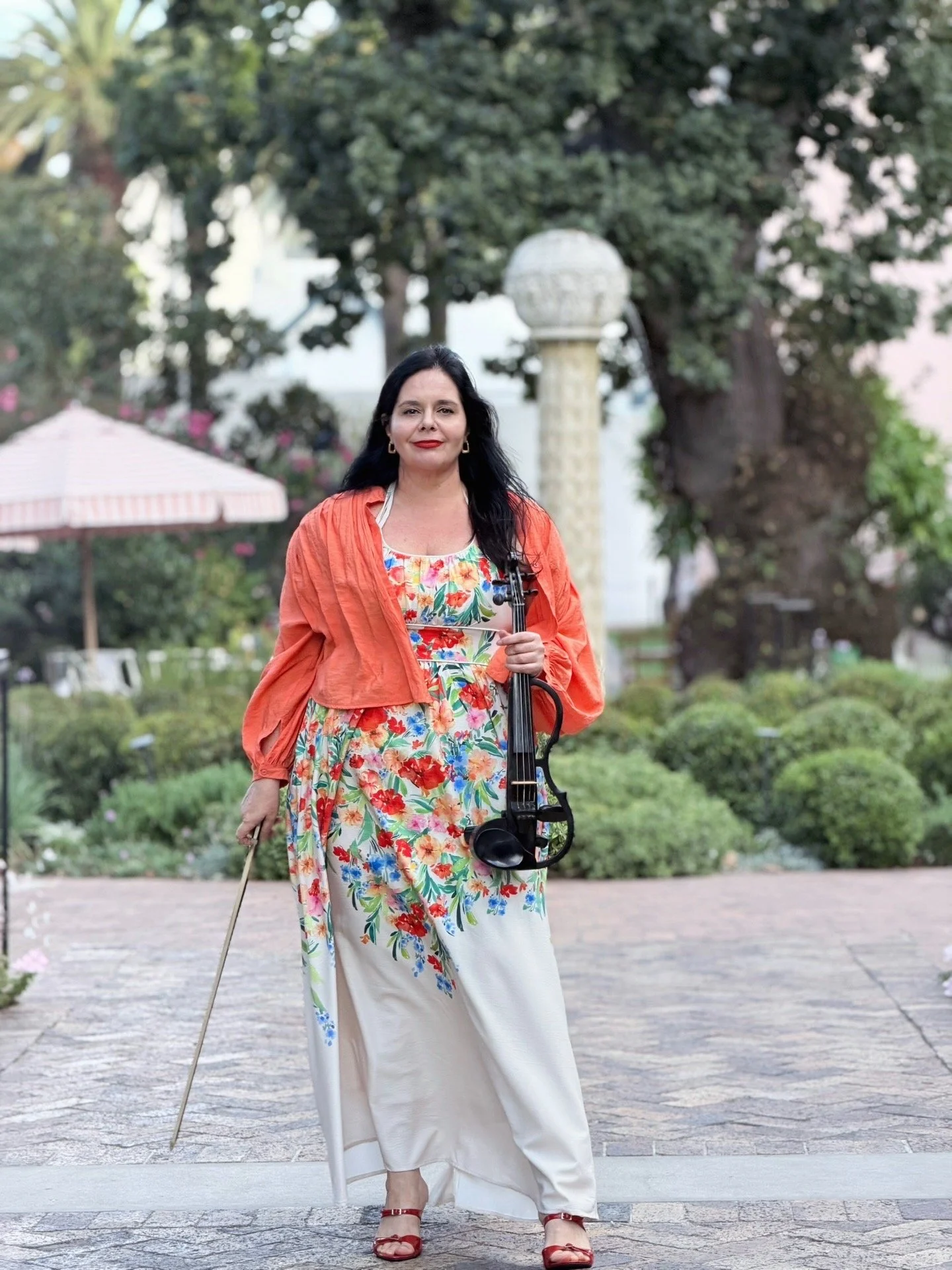 A woman walking outdoors on a paved pathway, holding a cello and a cane, wearing a colorful floral dress and an orange shawl, with a garden and large trees in the background.