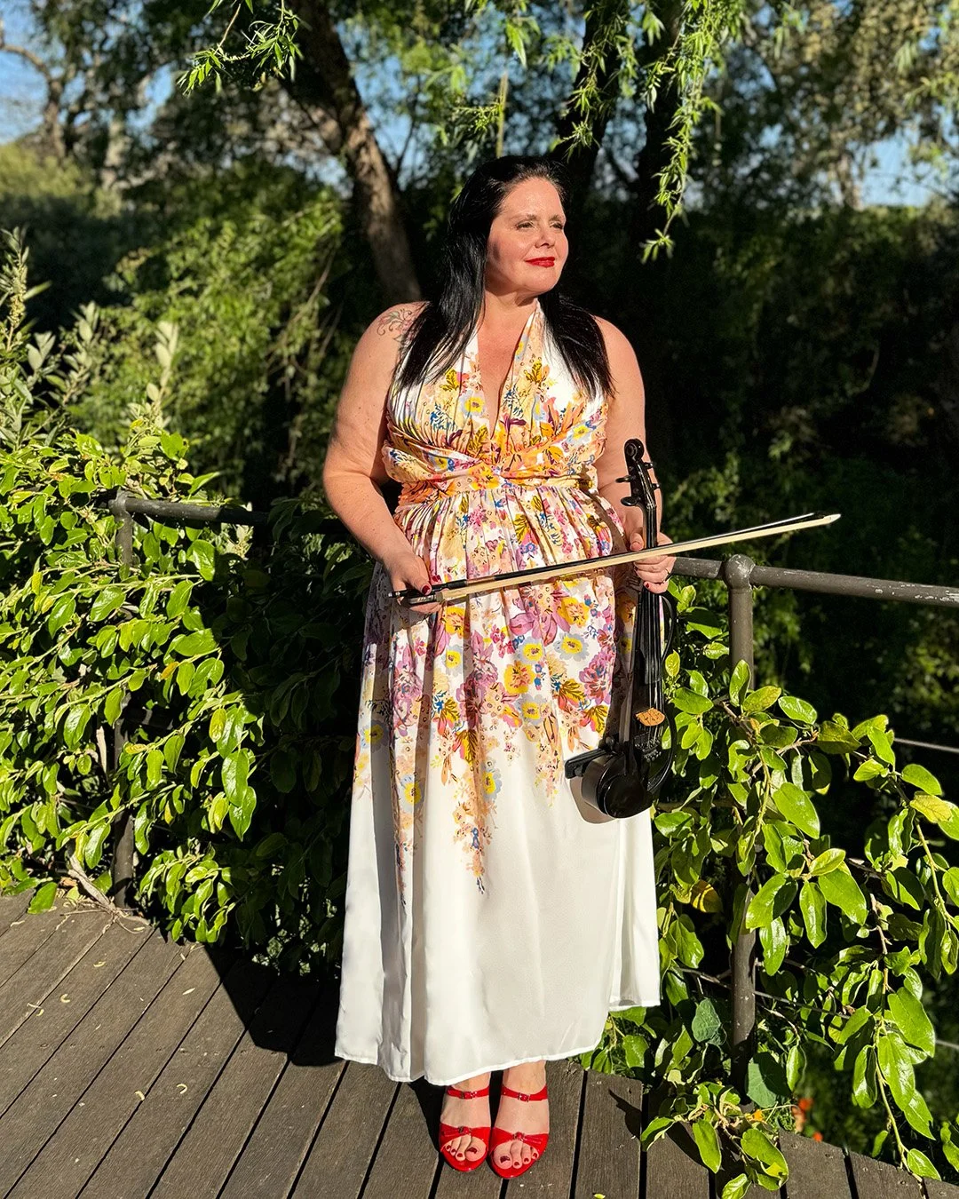 A woman with black hair and red lipstick stands on a wooden deck surrounded by greenery, wearing a floral dress with red high heels, holding a bass bow and a violin.