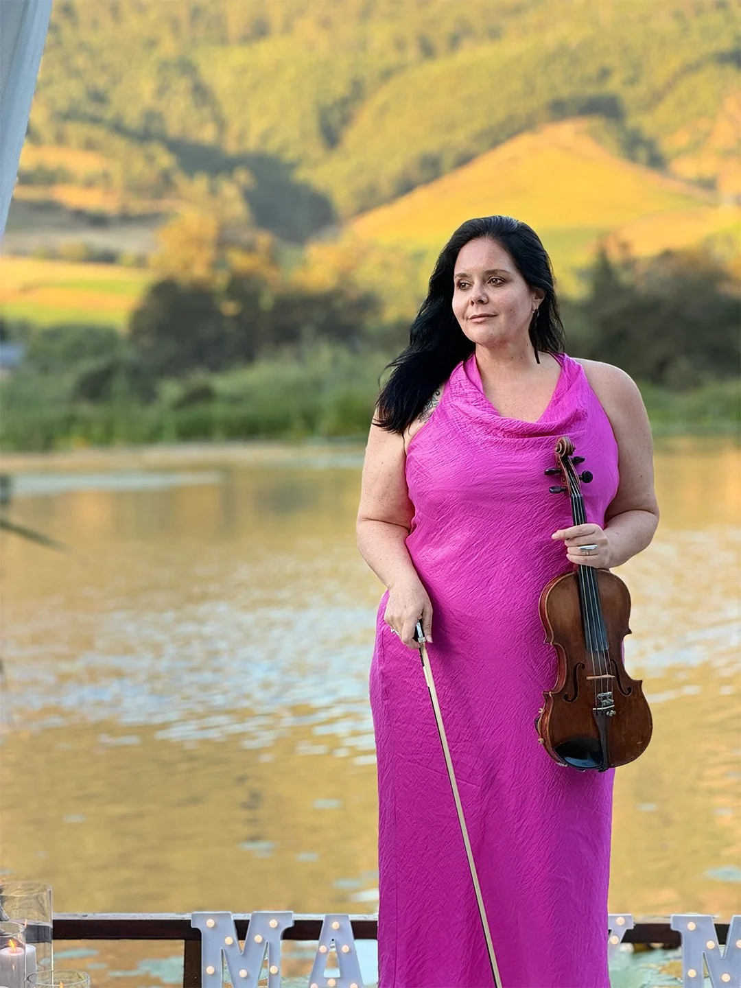 A woman with black hair playing a violin outdoors with mountains, a town, and a body of water in the background. There are flowers and a small box on a rock nearby.