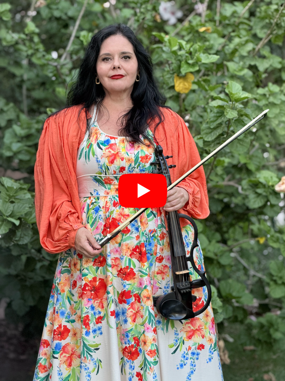 Woman with black hair holding a violin and a bow, wearing a colorful floral dress and an orange jacket, standing outdoors in front of green foliage.