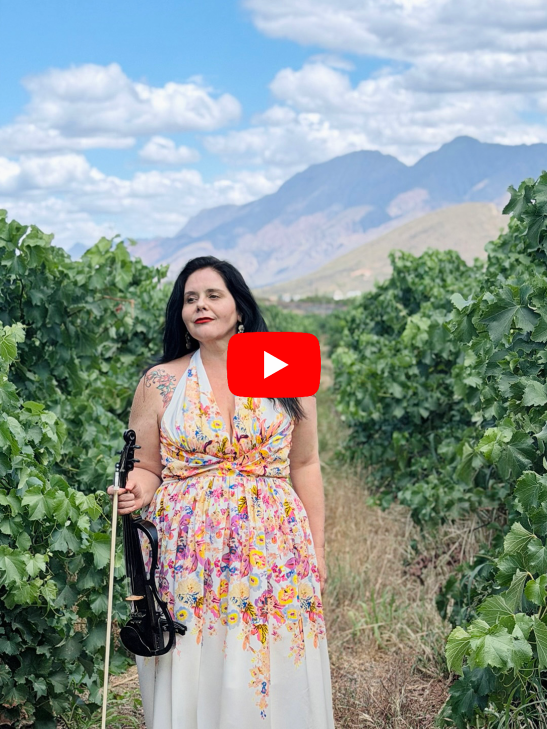 A woman with black hair wearing a colorful floral dress standing in a vineyard with green grapevines, mountains in the background, and a partly cloudy sky.