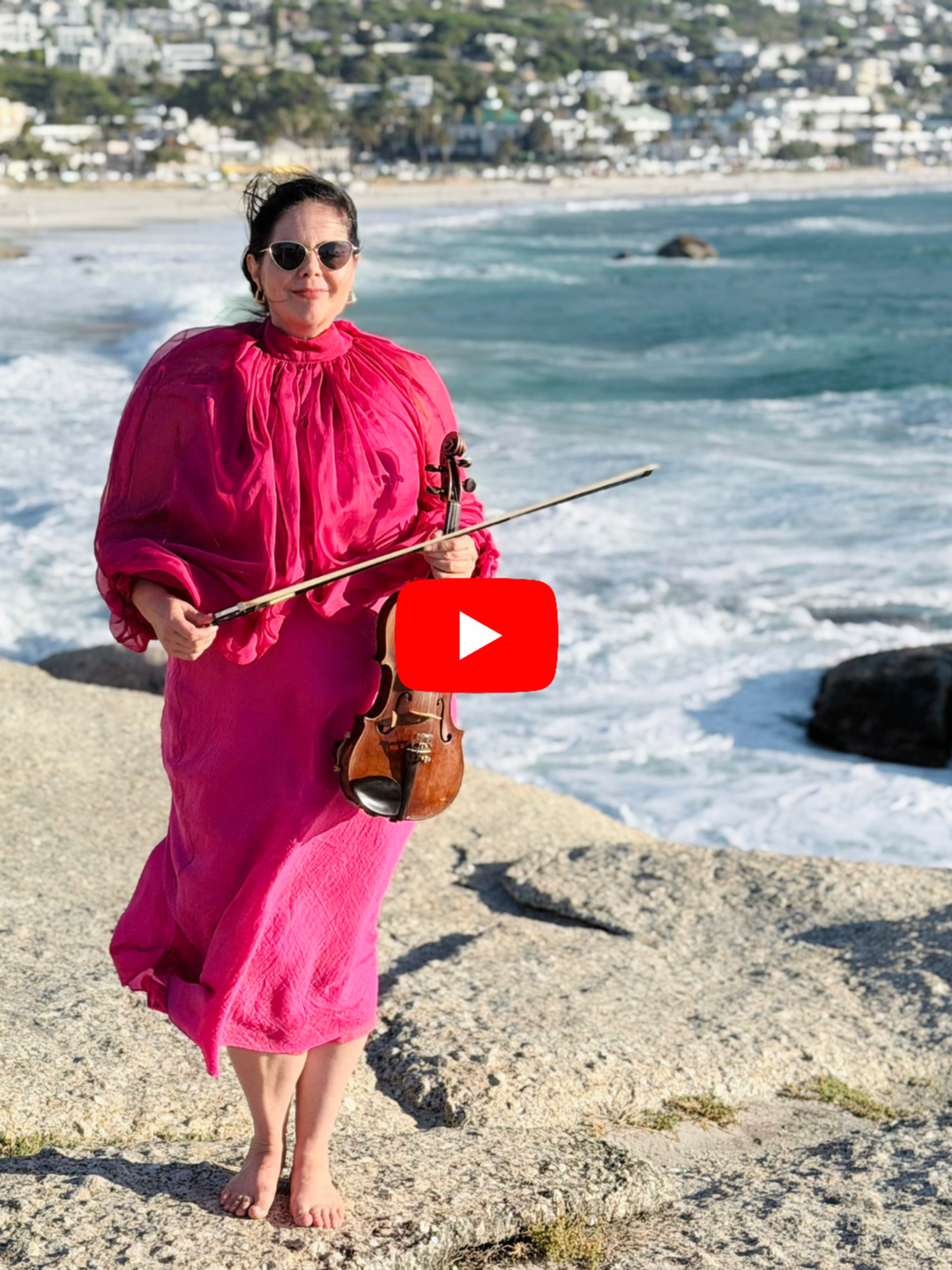 Woman standing barefoot on rocky beach, holding a violin and bow, wearing a bright pink dress and sunglasses, with ocean waves and houses in background.