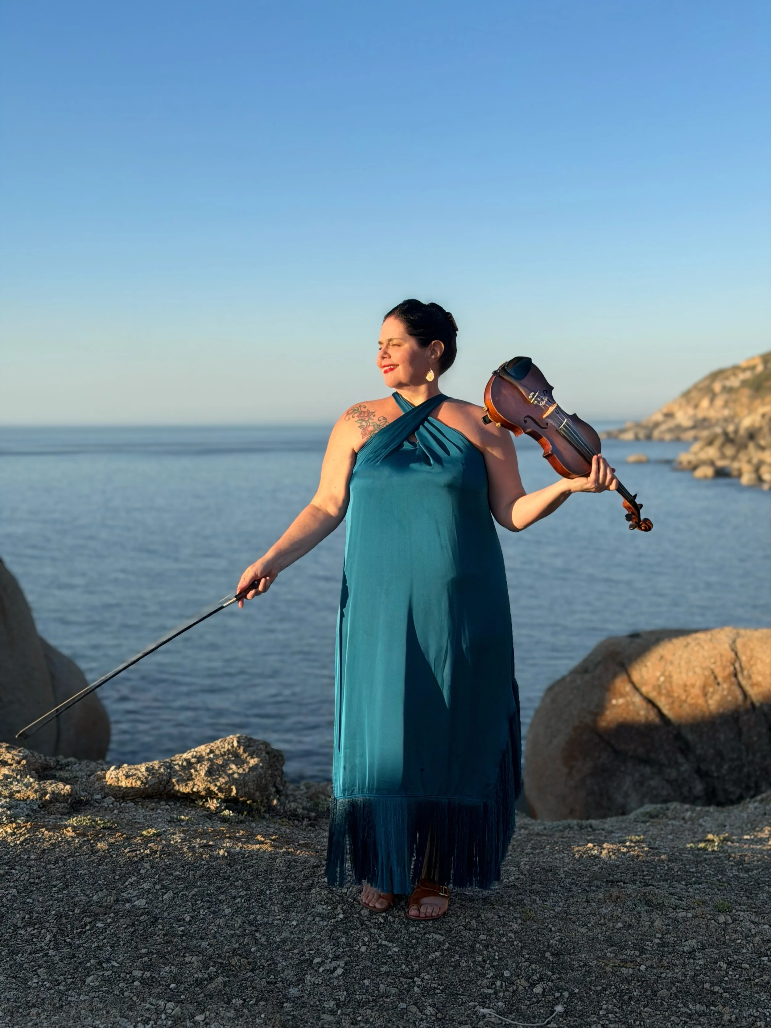 Woman with long dark hair in a teal dress holding a violin on a rocky beach with mountains and a coastal town in the background.