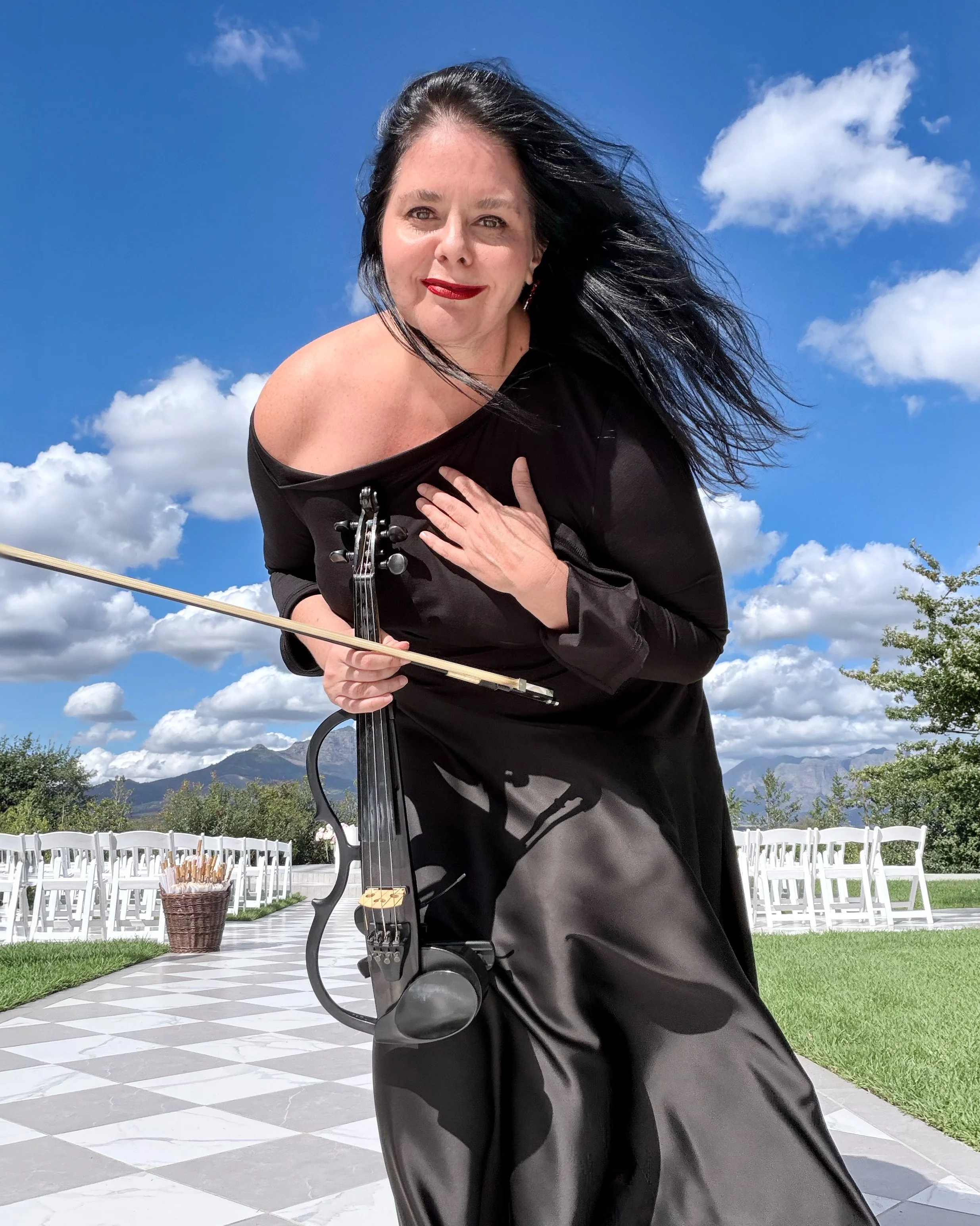 Larah Eksteen wearing a black dress, is posing with her hand on her chest and holding a black violin with her other hand, outdoors with a blue sky, white clouds, and mountains in the background.