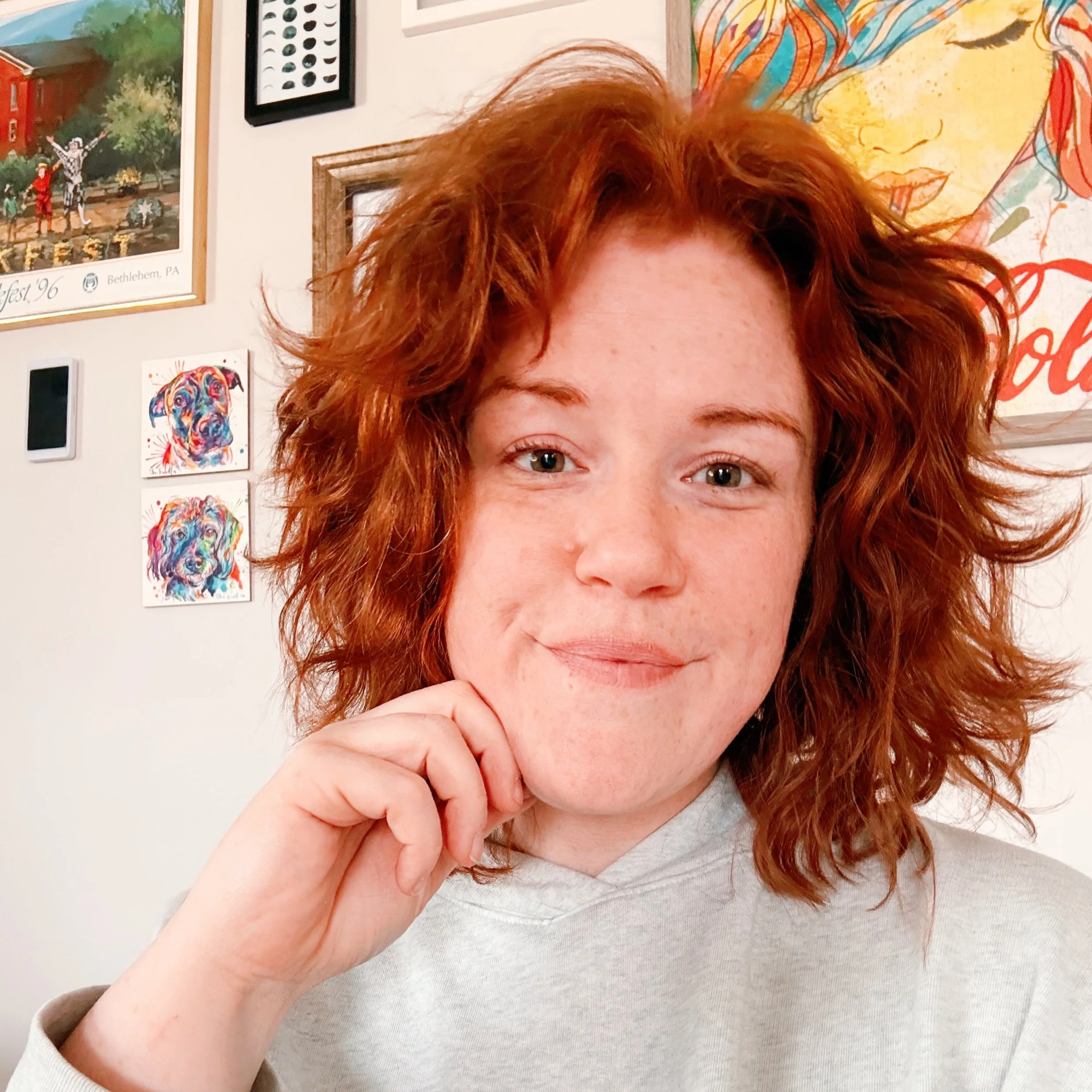 Close-up of a smiling woman with curly red hair, wearing a light gray hoodie, in a room with colorful artwork and framed pictures on the wall.
