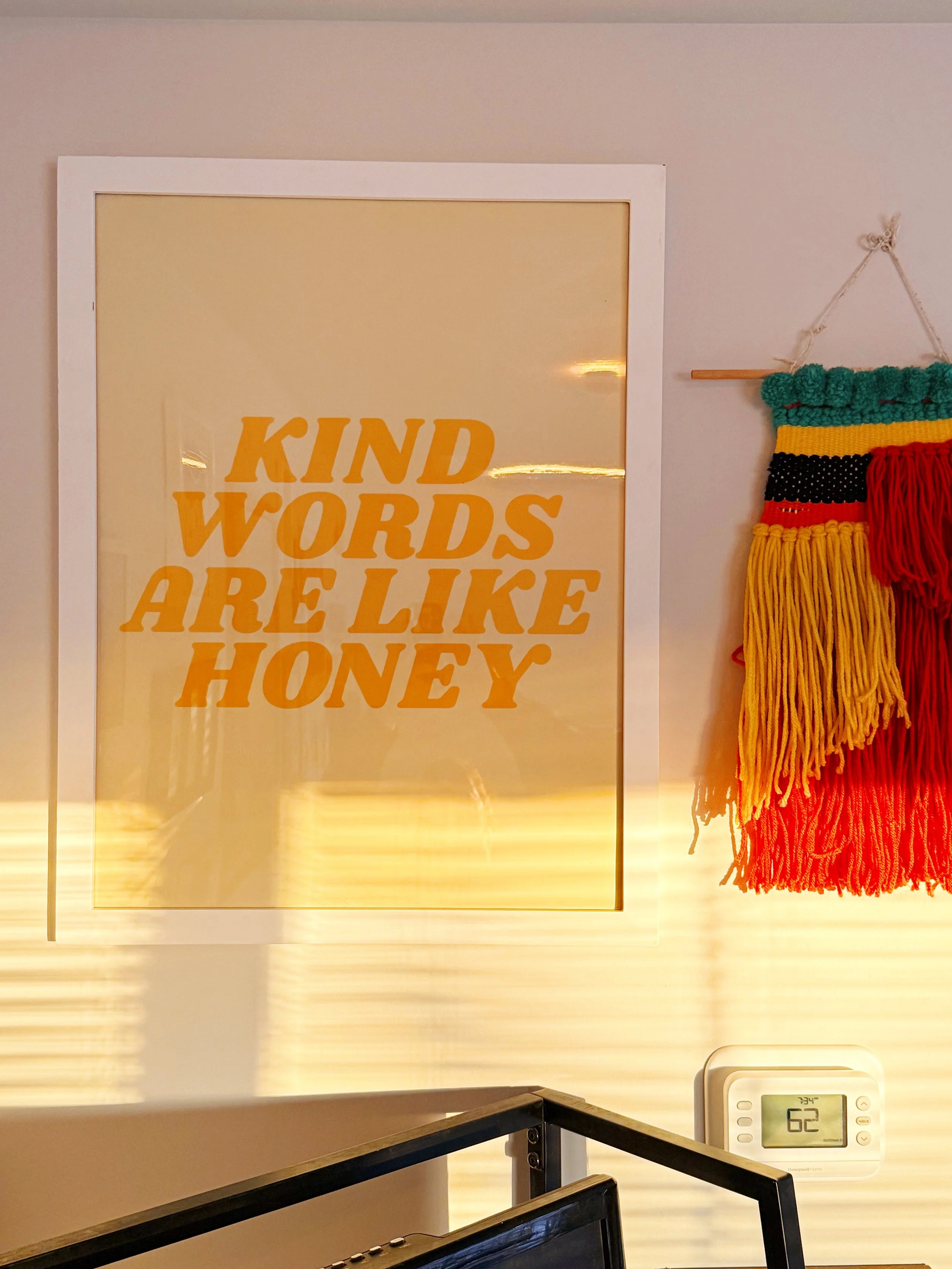 A framed sign that reads 'KIND WORDS ARE LIKE HONEY' in yellow text on a light background, with colorful yarn wall hanging to the right and a digital thermometer displaying 62 degrees below.
