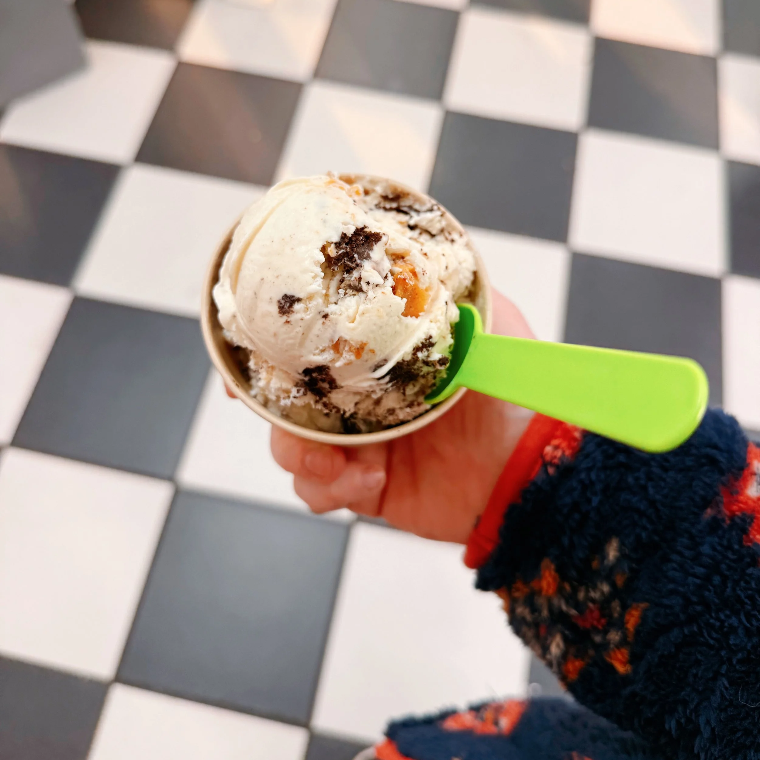 Hand holding a cup of cookies and cream ice cream with a green spoon, against a black and white checkered floor.