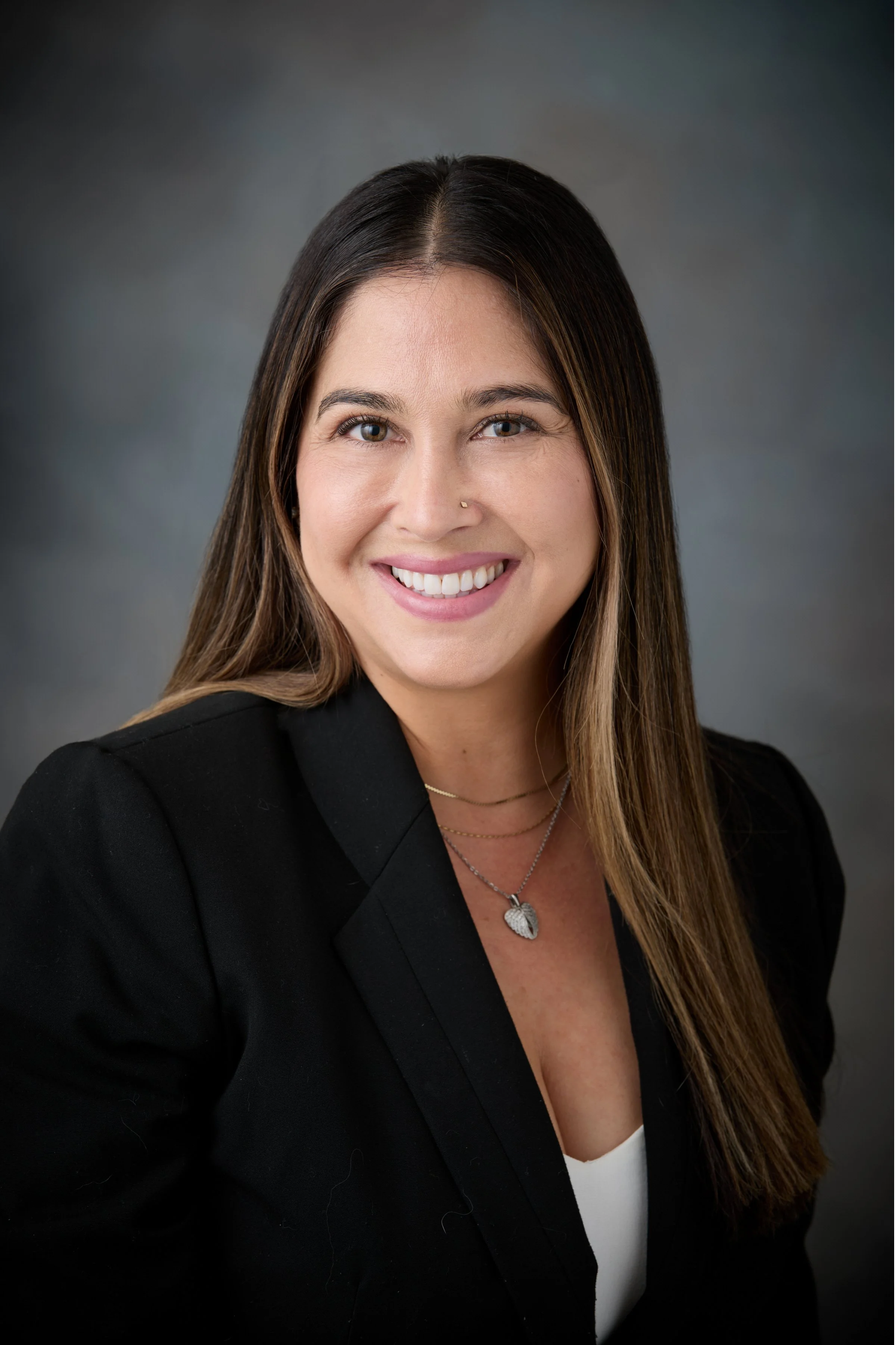 Professional headshot of a woman with long brown hair, smiling, wearing a black blazer and a white top, with a silver leaf-shaped necklace, against a dark gray background.
