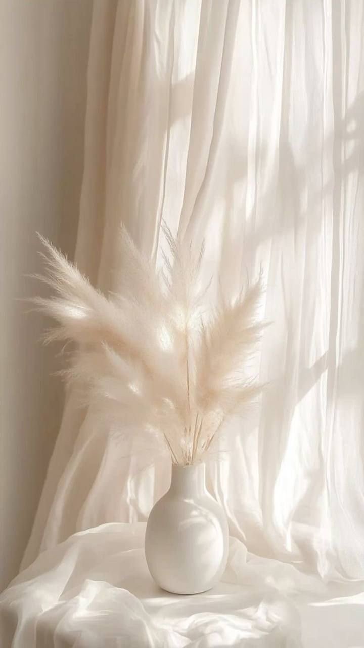 A white ceramic vase with dried pampas grass on a white cloth, in front of a sheer white curtain.