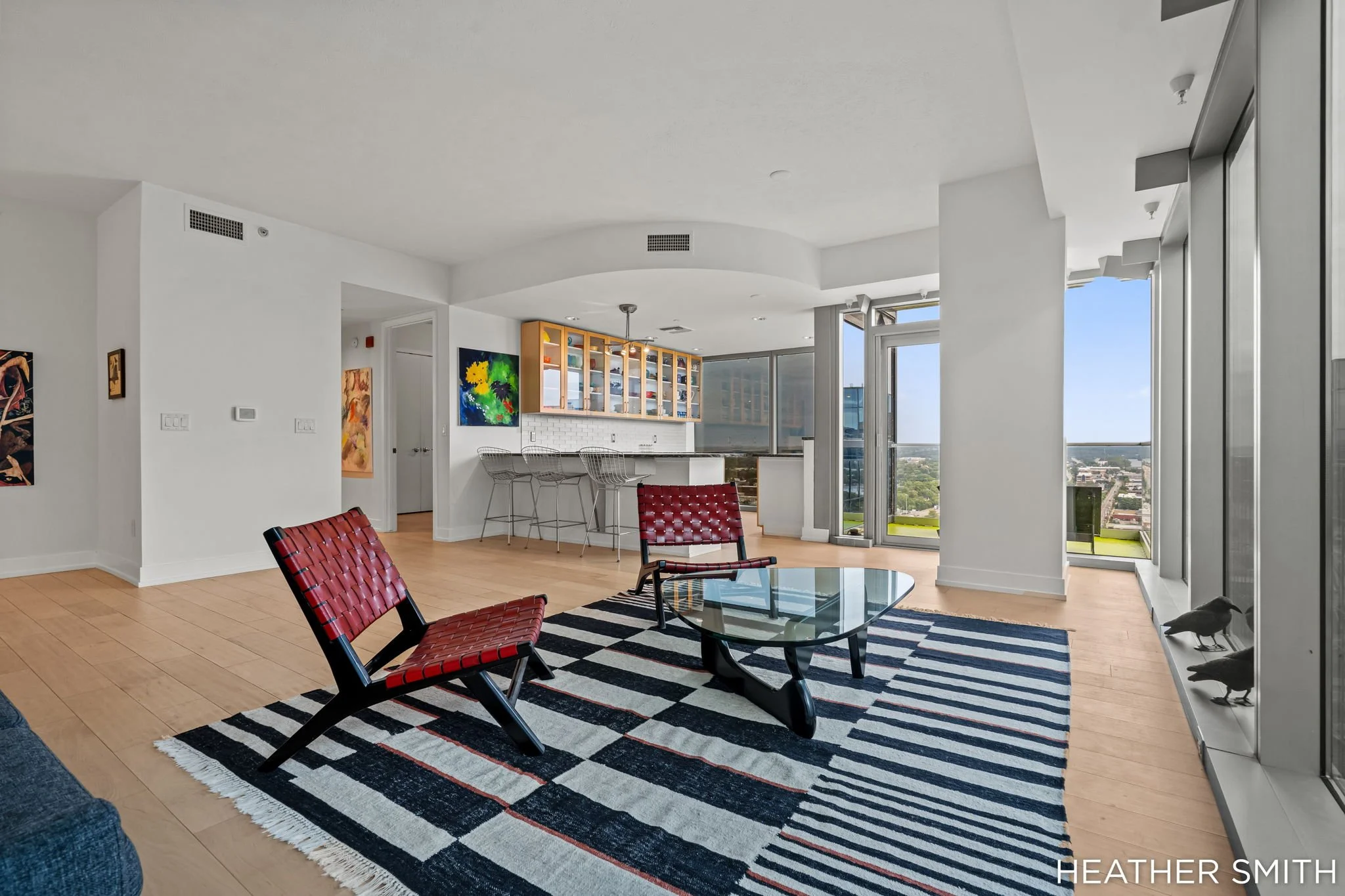 Modern apartment living room with large windows, red woven chairs, glass coffee table on a black-and-white striped rug, wall art, and a kitchen area with barstools, open shelves, and city views.