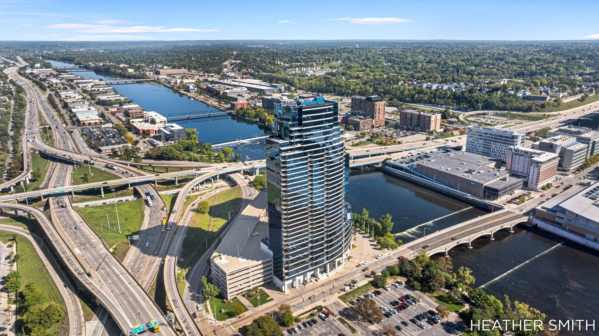 Aerial view of a city showing highways, bridges, a river, and tall buildings surrounded by green trees under a partly cloudy sky.