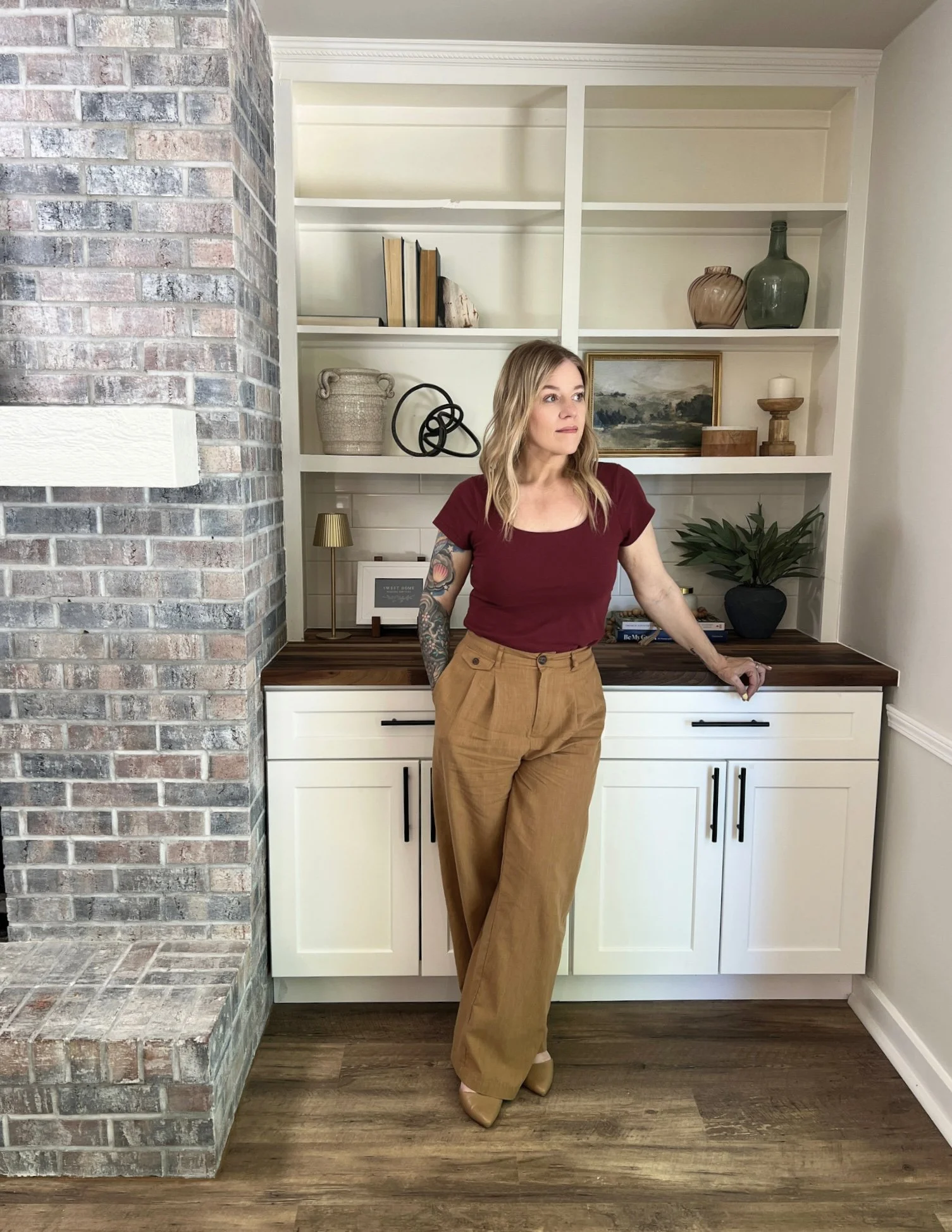 A woman with blonde hair, wearing a burgundy top and khaki wide-legged pants, leaning against a kitchen counter with built-in white cabinets and shelves, decorated with books, vases, a painting, and a candle holder.