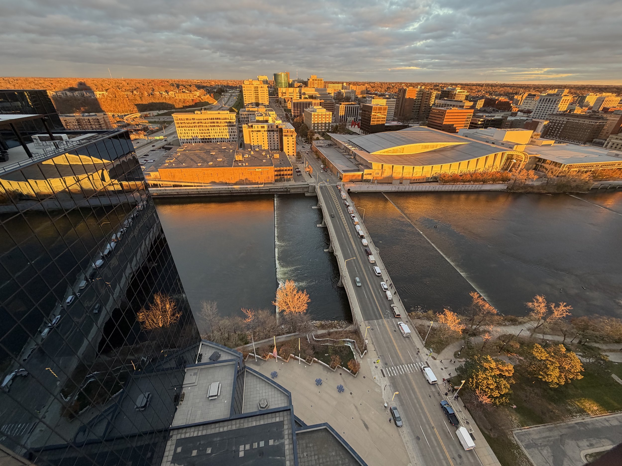 A cityscape at sunset with a river in the foreground, a bridge crossing the river, and modern buildings with glass and concrete facades illuminated by the golden light, with a mix of residential and commercial structures, under a partly cloudy sky.