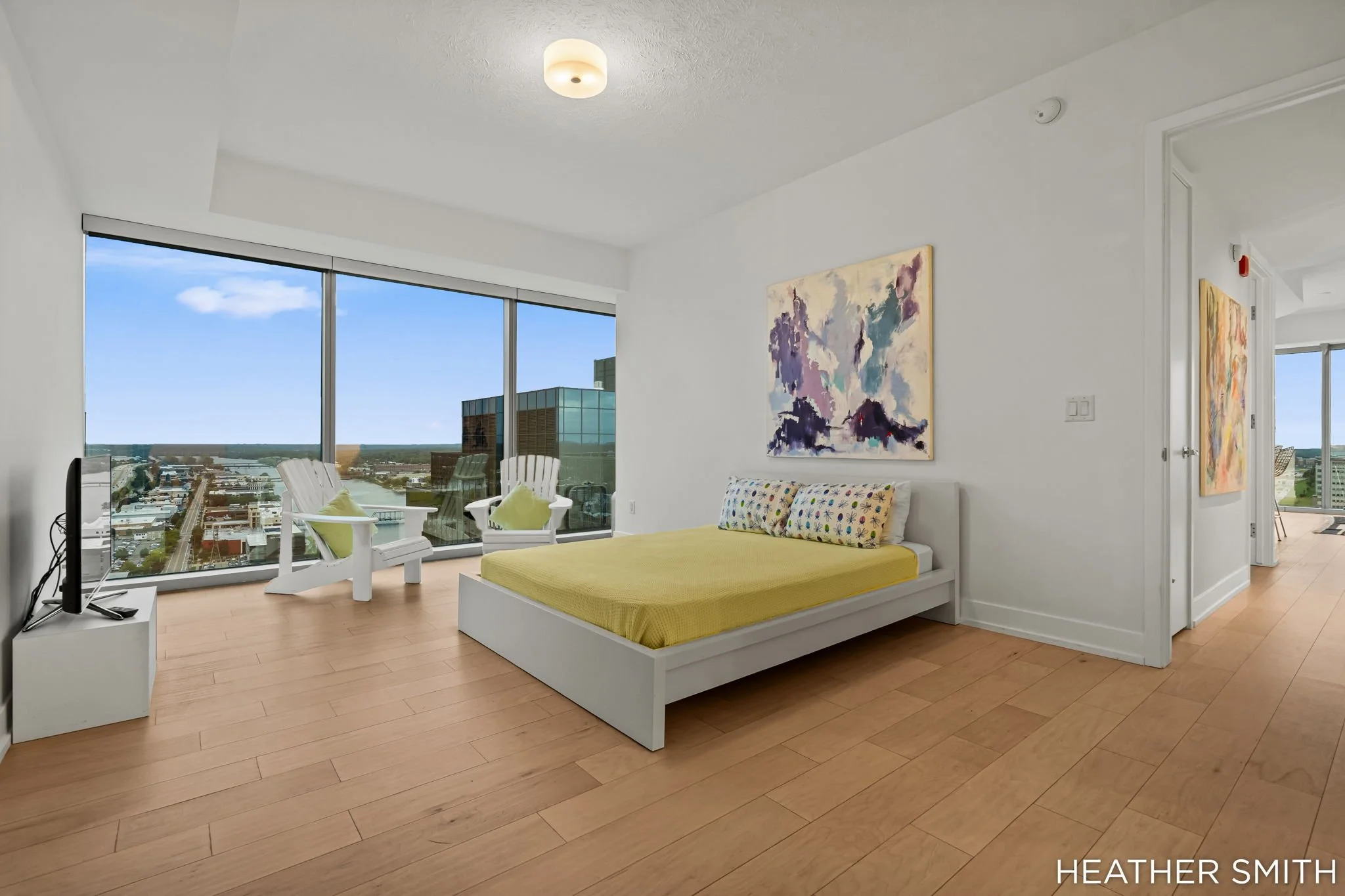 Interior of a modern apartment bedroom with large windows showing a city view, a bed with yellow bedding and patterned pillows, abstract wall art, and a TV on a white stand.