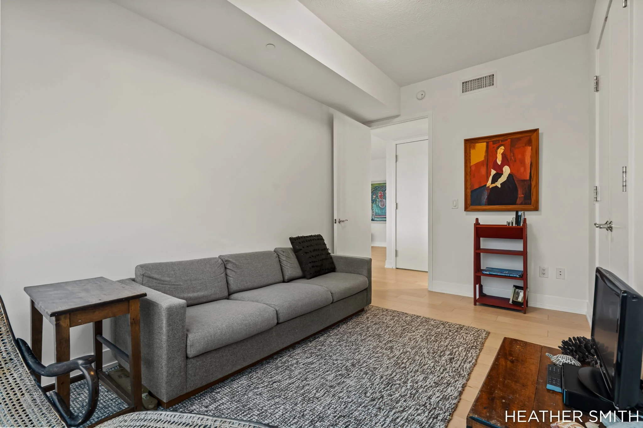Living room with gray sofa, wooden side table, black decorative pillows, area rug, white walls, framed art, red bookshelves, and a television.