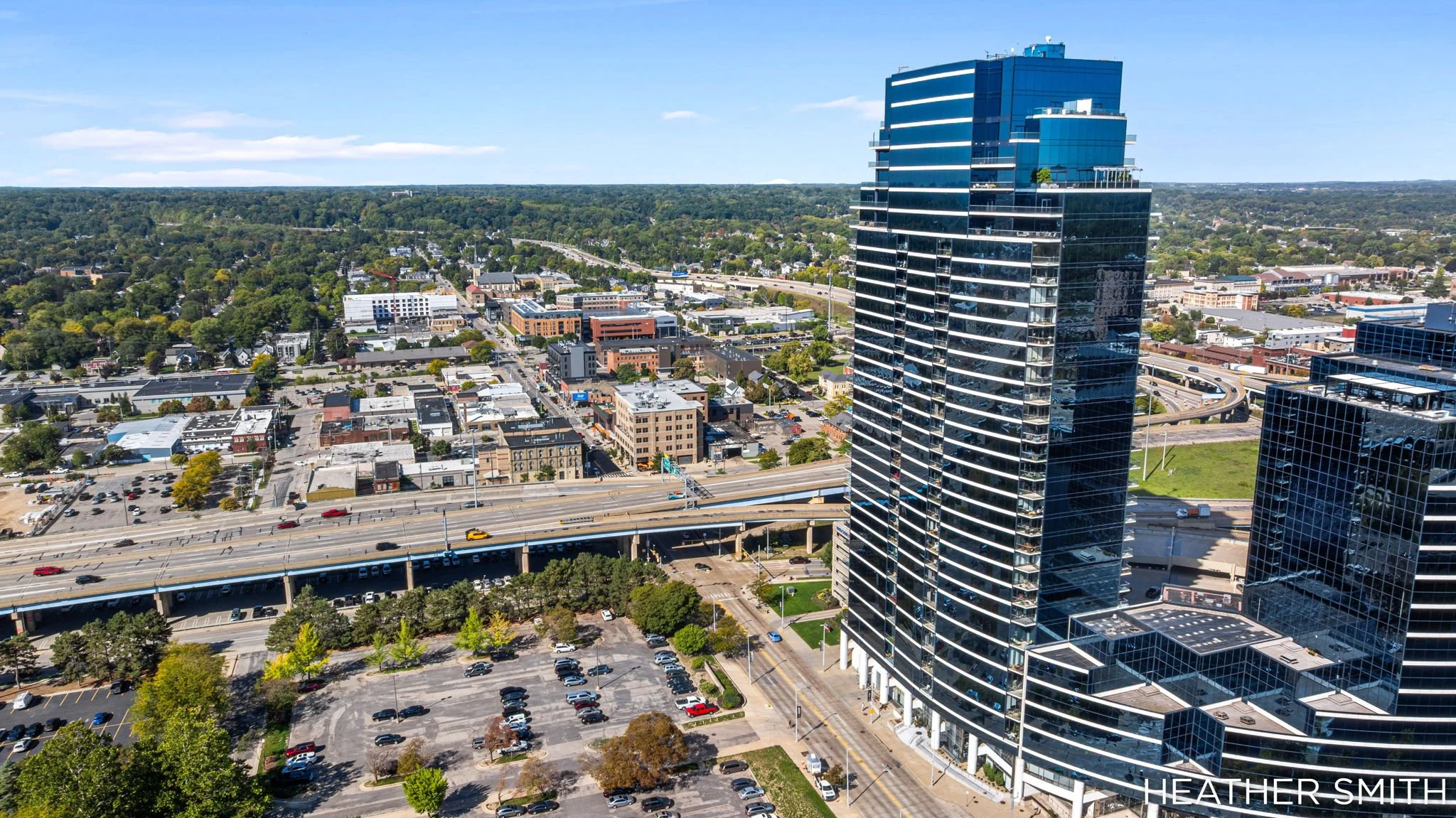 Aerial view of a cityscape with a tall modern glass skyscraper in the foreground, smaller buildings, parking lots, and a highway surrounded by greenery under a blue sky.