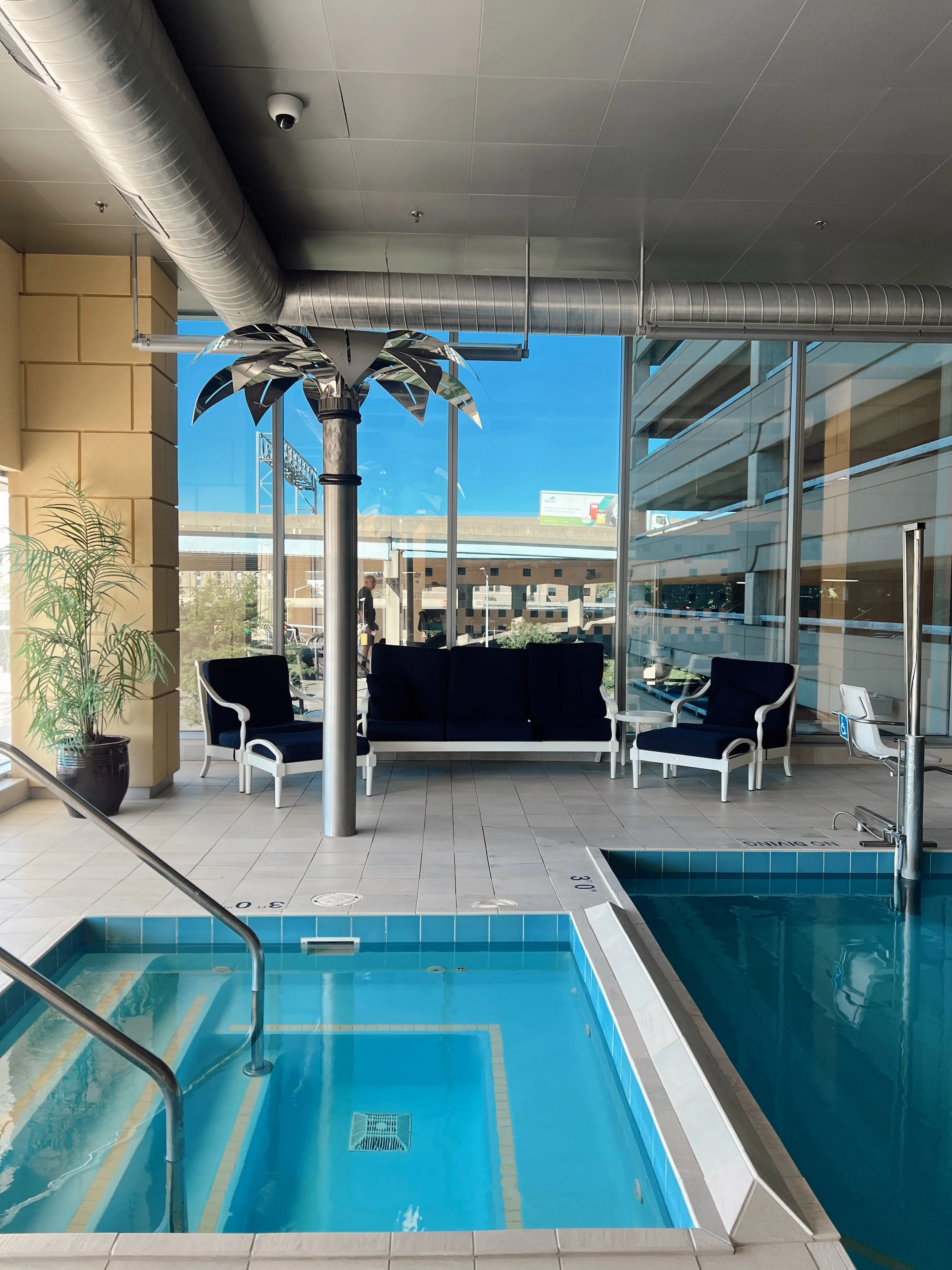 Indoor swimming pool with blue water, starting block, metal handrail, black chairs, potted plant, glass walls, and reflection of city buildings outside.