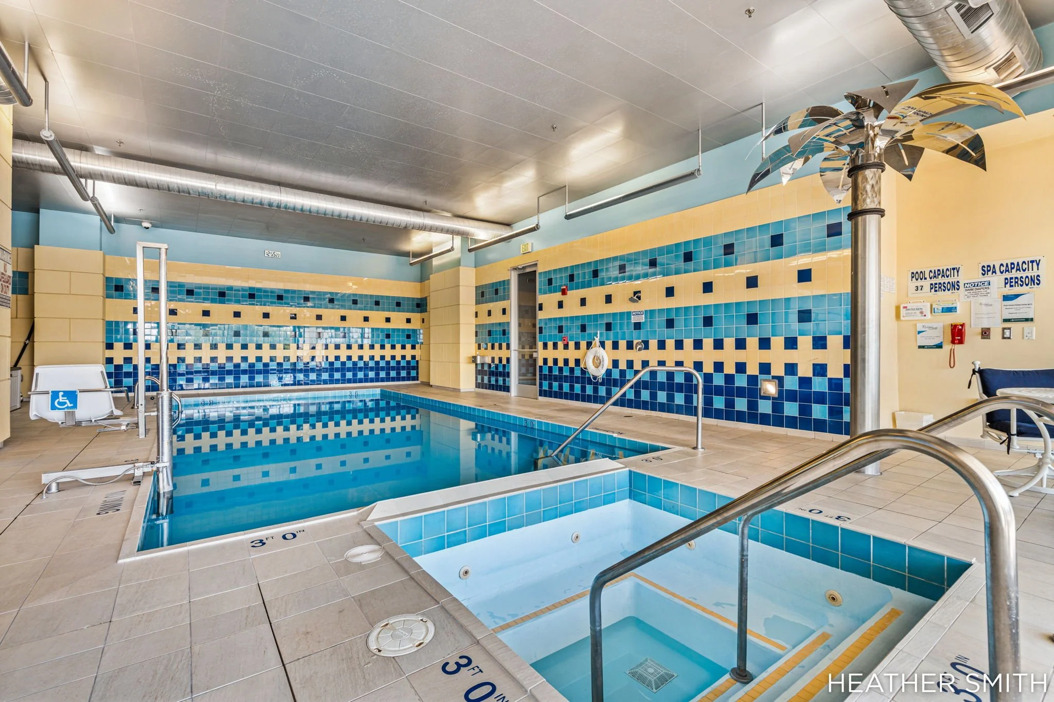Indoor swimming pool with blue and yellow tiled walls, a small pool with a lift for accessibility, pool and spa capacity signs, and a decorative metallic palm tree sculpture.
