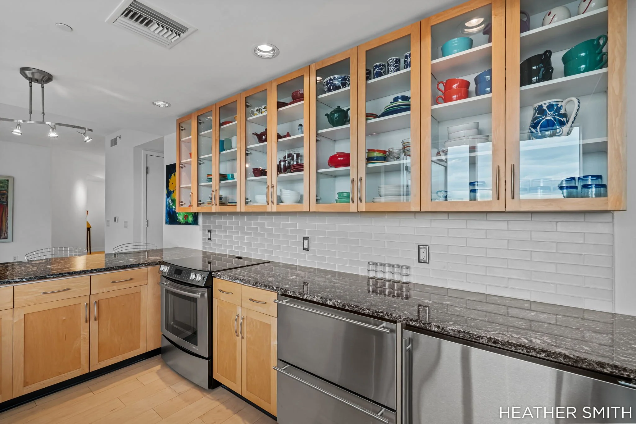 Kitchen with wooden cabinets displaying colorful dishes, granite countertops, stainless steel appliances, and a white brick backsplash.