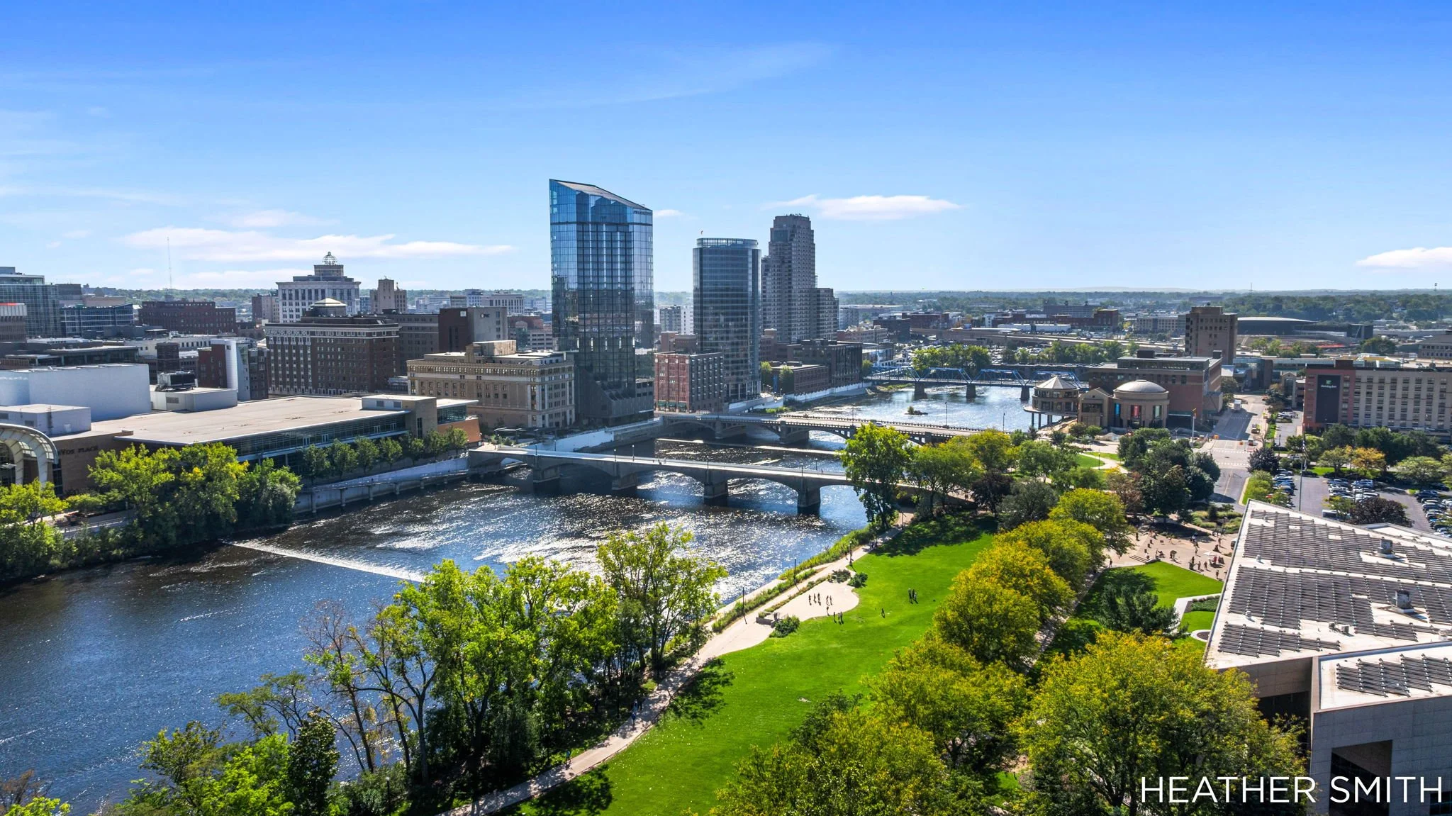 A cityscape viewed from an elevated position showing a river, green park, and tall modern buildings under a blue sky.