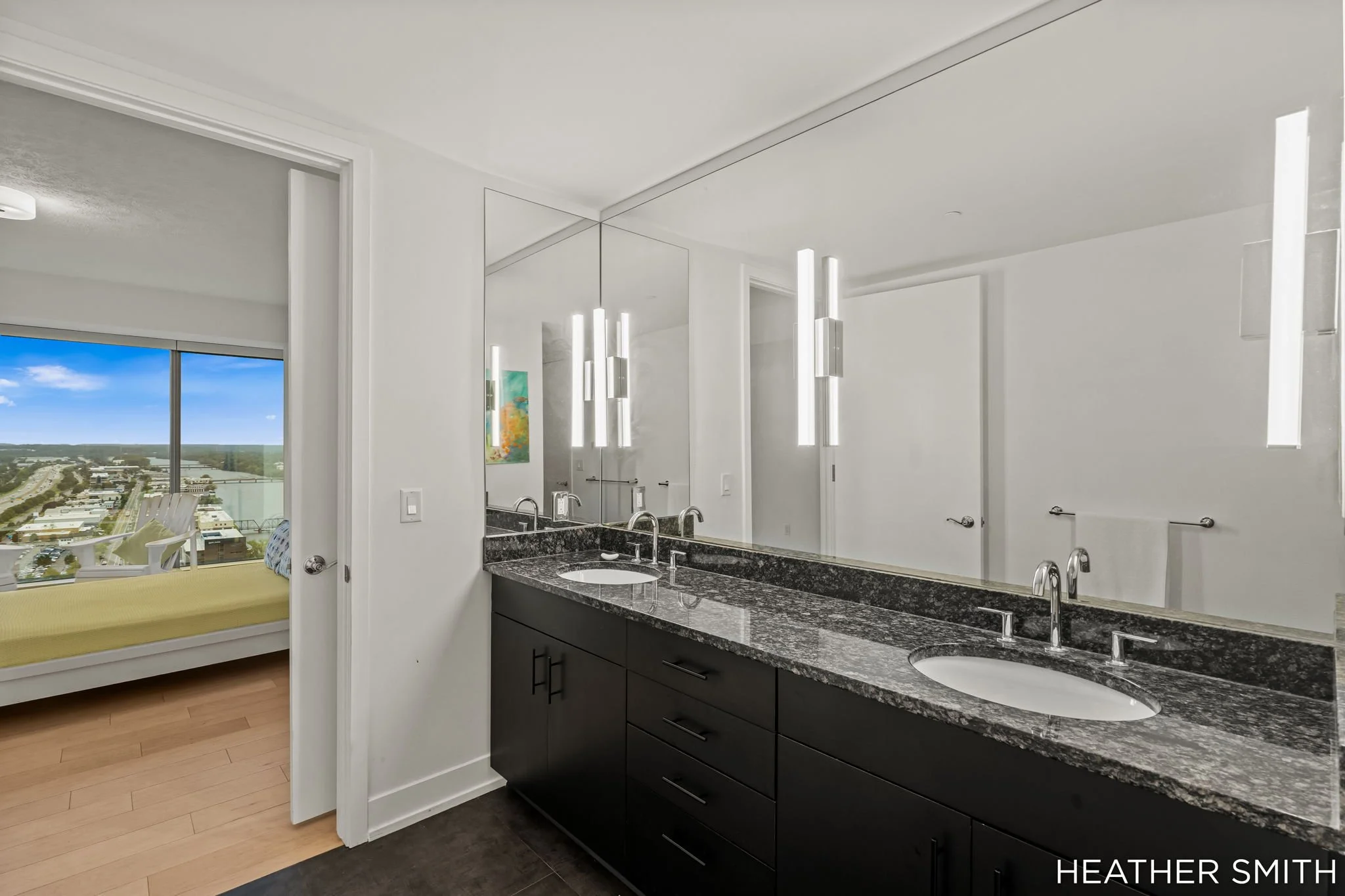Modern bathroom with black granite countertop, white walls, and a large mirror with vertical lights. Visible window shows a city view and a bedroom with a yellow bed.