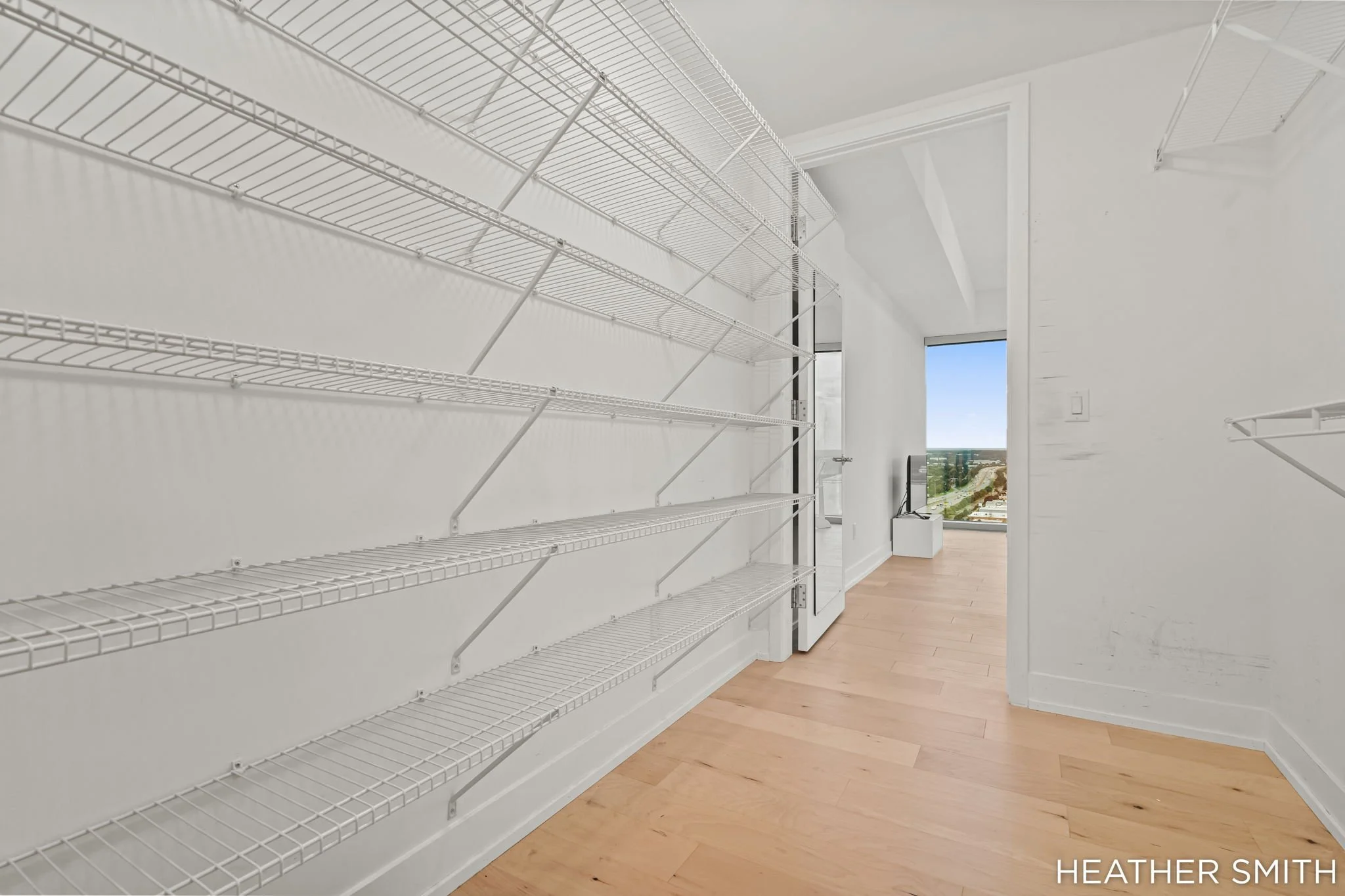 Empty white wire shelves mounted along a white wall in a room with hardwood floors and an open door leading to a balcony view.