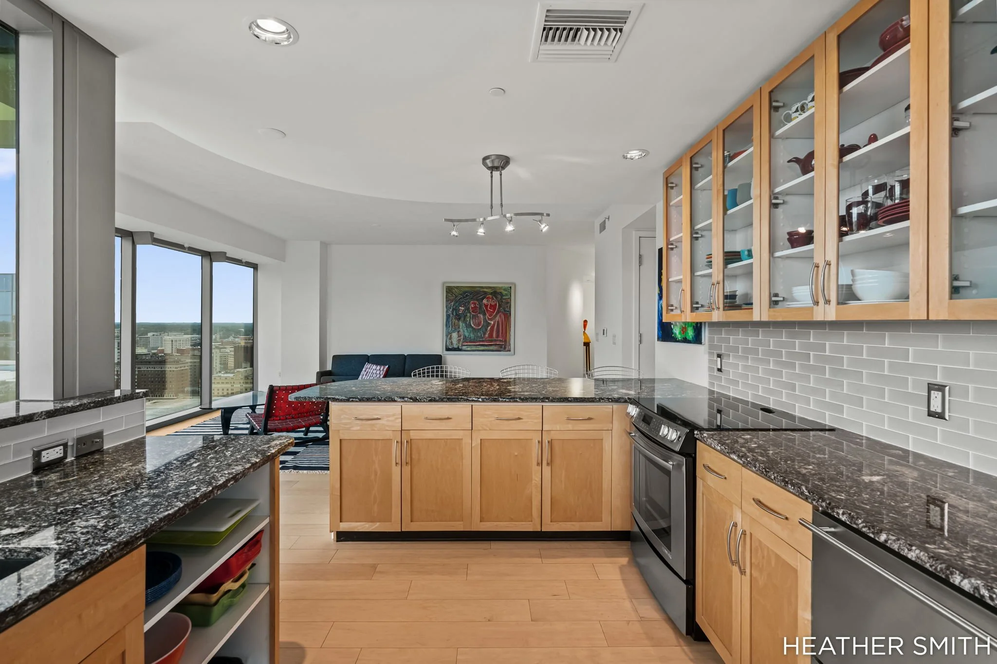 Modern kitchen with granite countertops, light wooden cabinets with glass doors, and stainless steel appliances. There's a view of city buildings outside large windows, with artwork on the walls and a small dining area visible.
