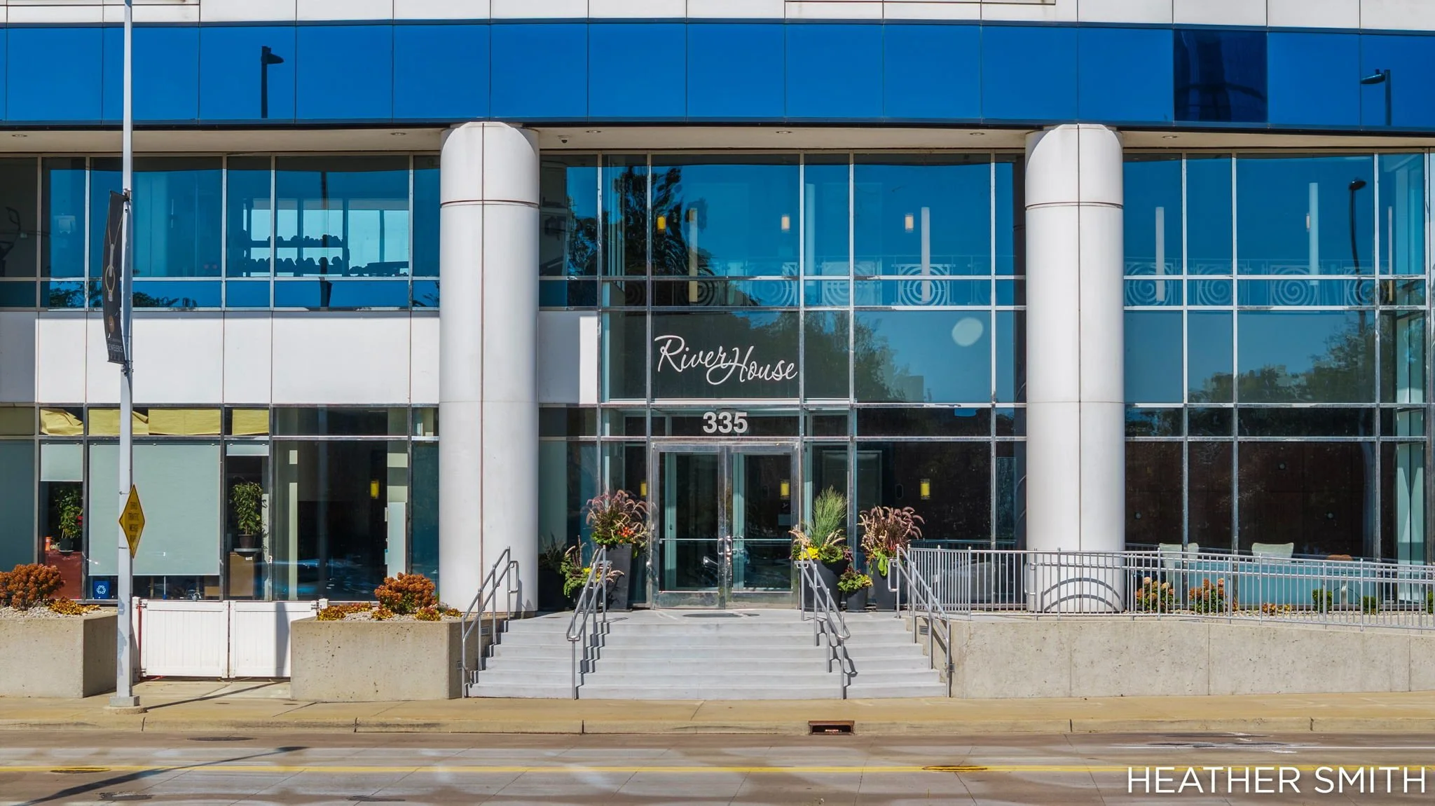 Front view of a modern glass building named Riverside. The entrance has steps, potted plants, and glass doors. Reflective windows show trees and sky.