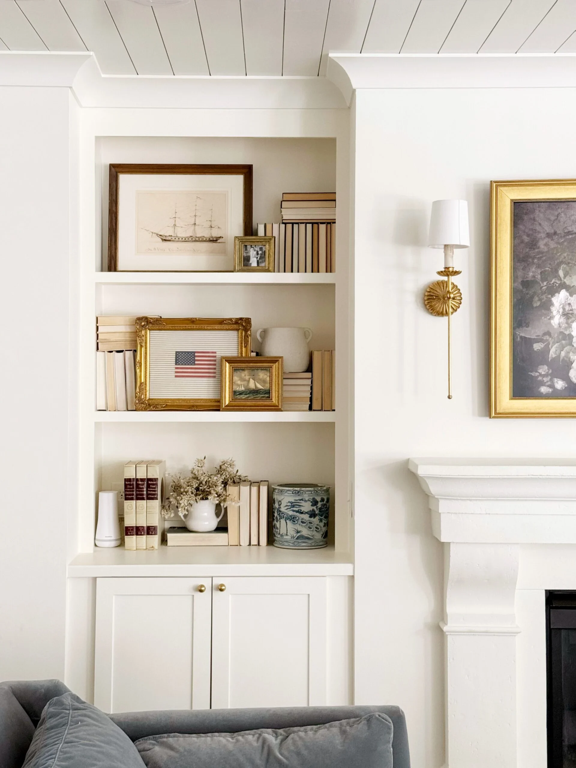 A built-in white bookshelf filled with framed pictures, books, a white vase, and decorative items. A vintage painting of a ship is on top, with other framed photos and books arranged around. To the right, there is a white wall with a gold wall sconce and a dark fireplace with a white mantle.