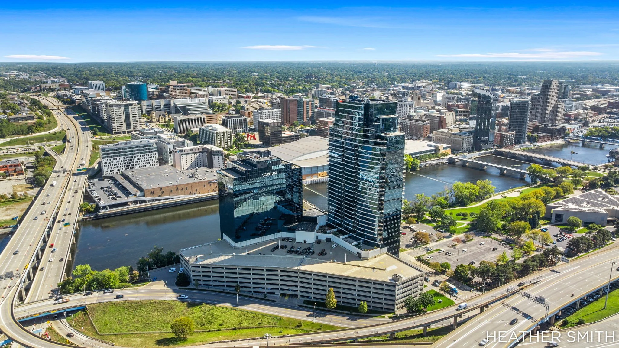 Aerial view of a city with tall buildings, bridges over a river, and green parks. Wide highway with cars runs through the scene on a sunny day with blue sky.