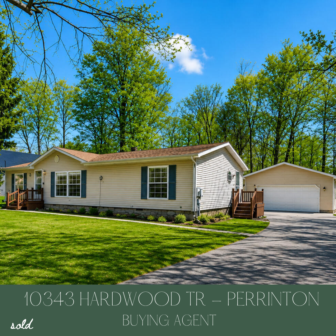 A single-story house with beige siding, green shutters, and a small front porch, surrounded by green grass and tall trees, under a blue sky.